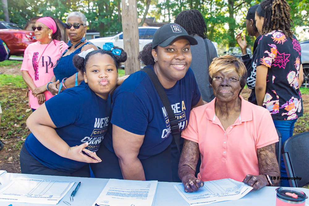 Group of people outdoors, including two women and a girl, smiling and sitting at a table with papers, trees in background.