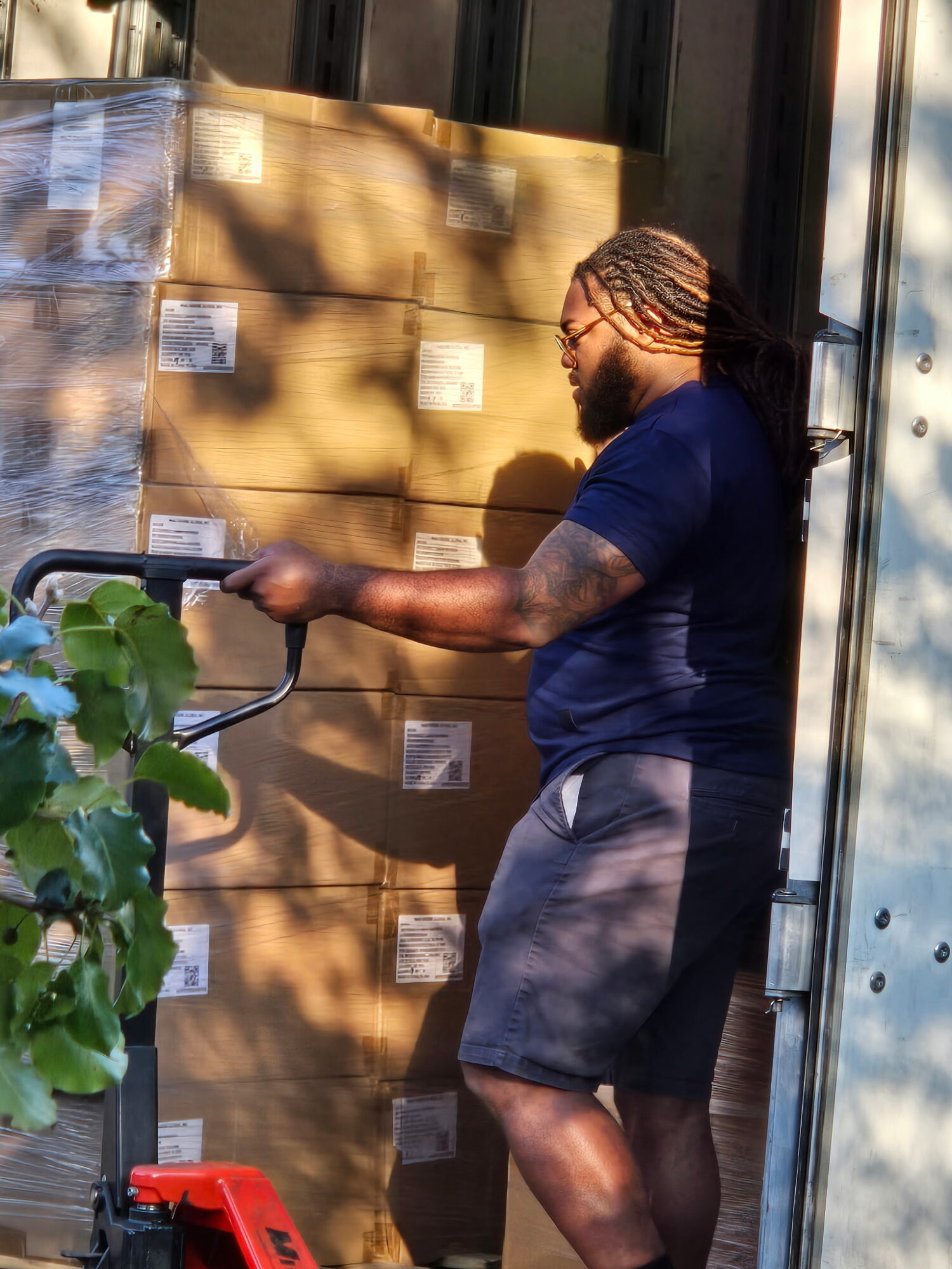 Man with braided hair and glasses stands next to stacked boxes outdoors, holding a clipboard, with sunlight and shadows on the scene.