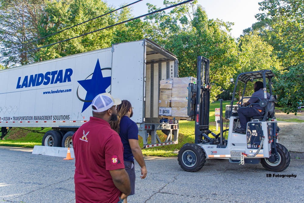 People unloading boxes from a truck using a forklift outdoors with trees in the background.