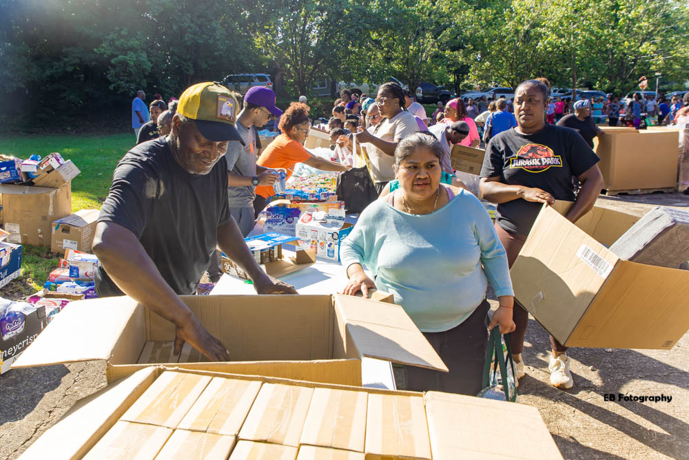 People gather outdoors around tables with boxes and supplies, some smiling and interacting, in a sunny park setting.