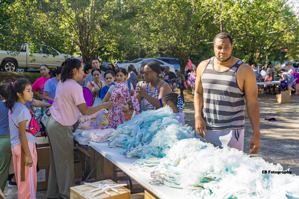 Group of children and adults gathered around a table with blue and white cotton candy outdoors, trees and parked cars in background.