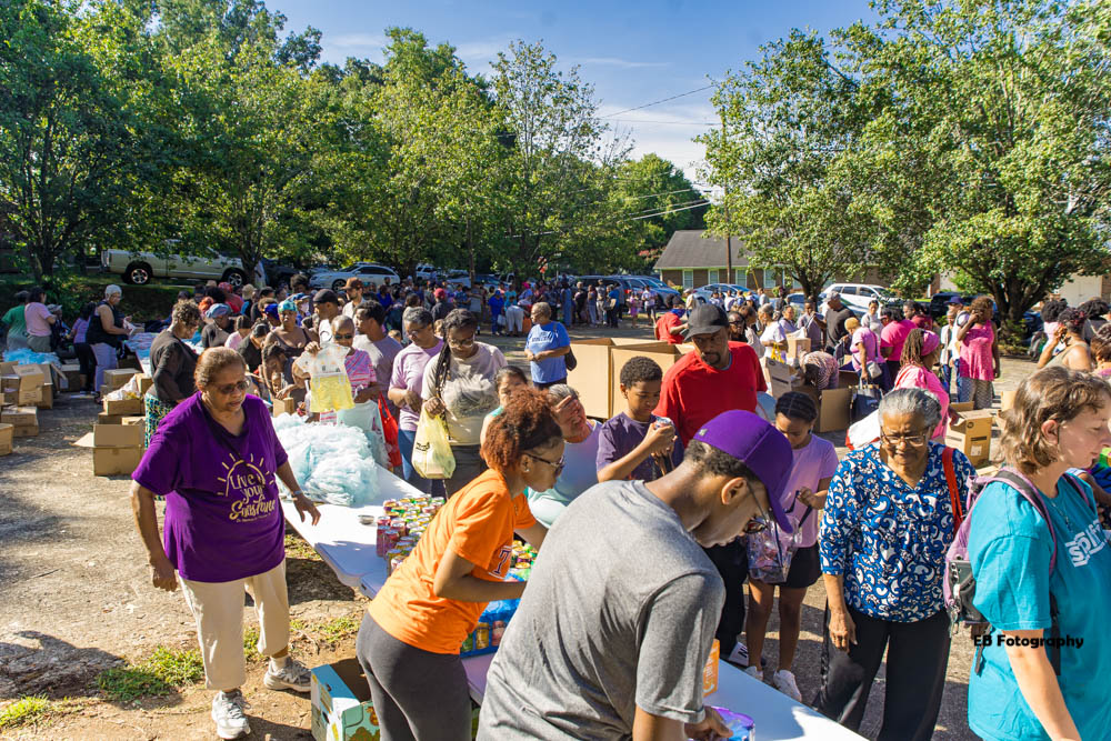 Crowd of people gathered outdoors in a park with trees and a small building in the background.