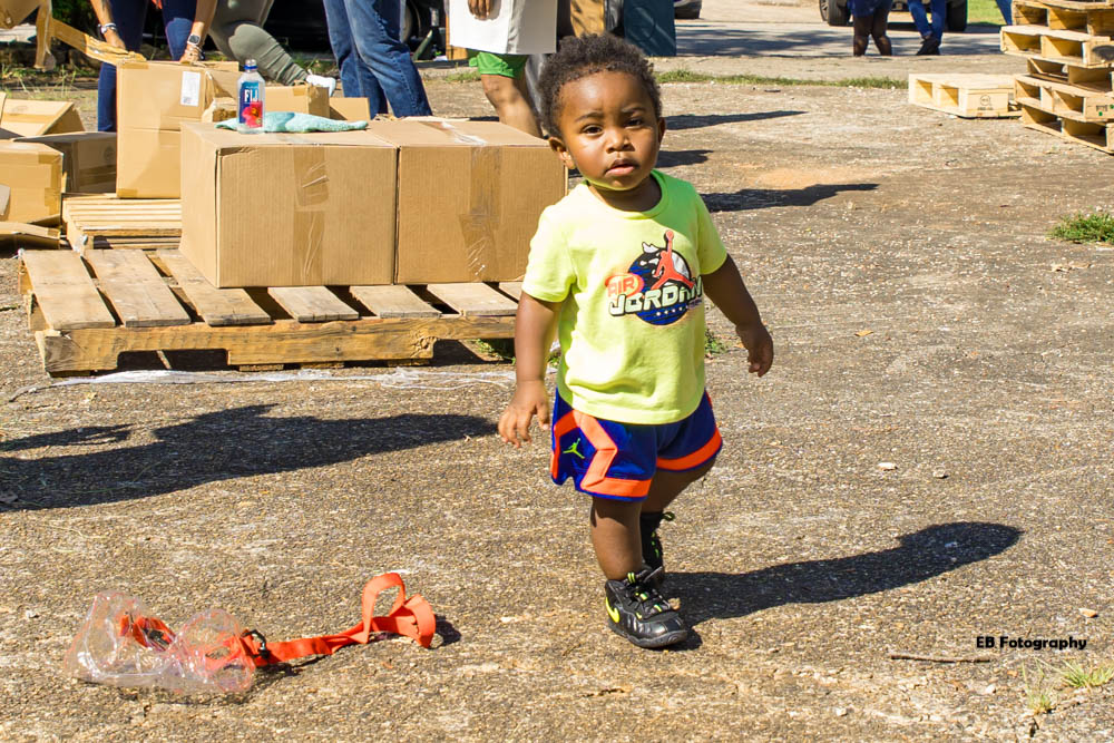 Young child with curly hair wearing a yellow shirt and blue shorts walking outdoors on a dirt surface.