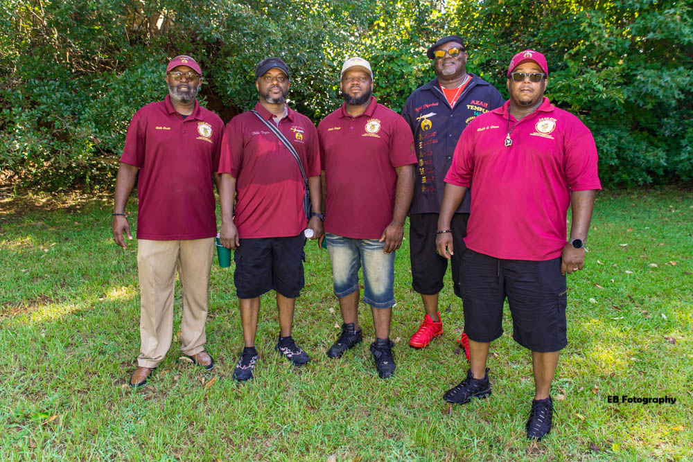 Five people stand outdoors on grass with trees in the background, wearing maroon and dark blue sports shirts and caps.