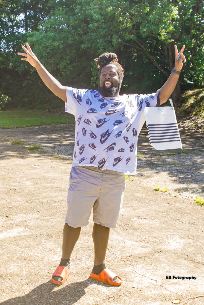 Man with arms raised outdoors, smiling, wearing a patterned shirt, shorts, and orange sandals, with trees in background.