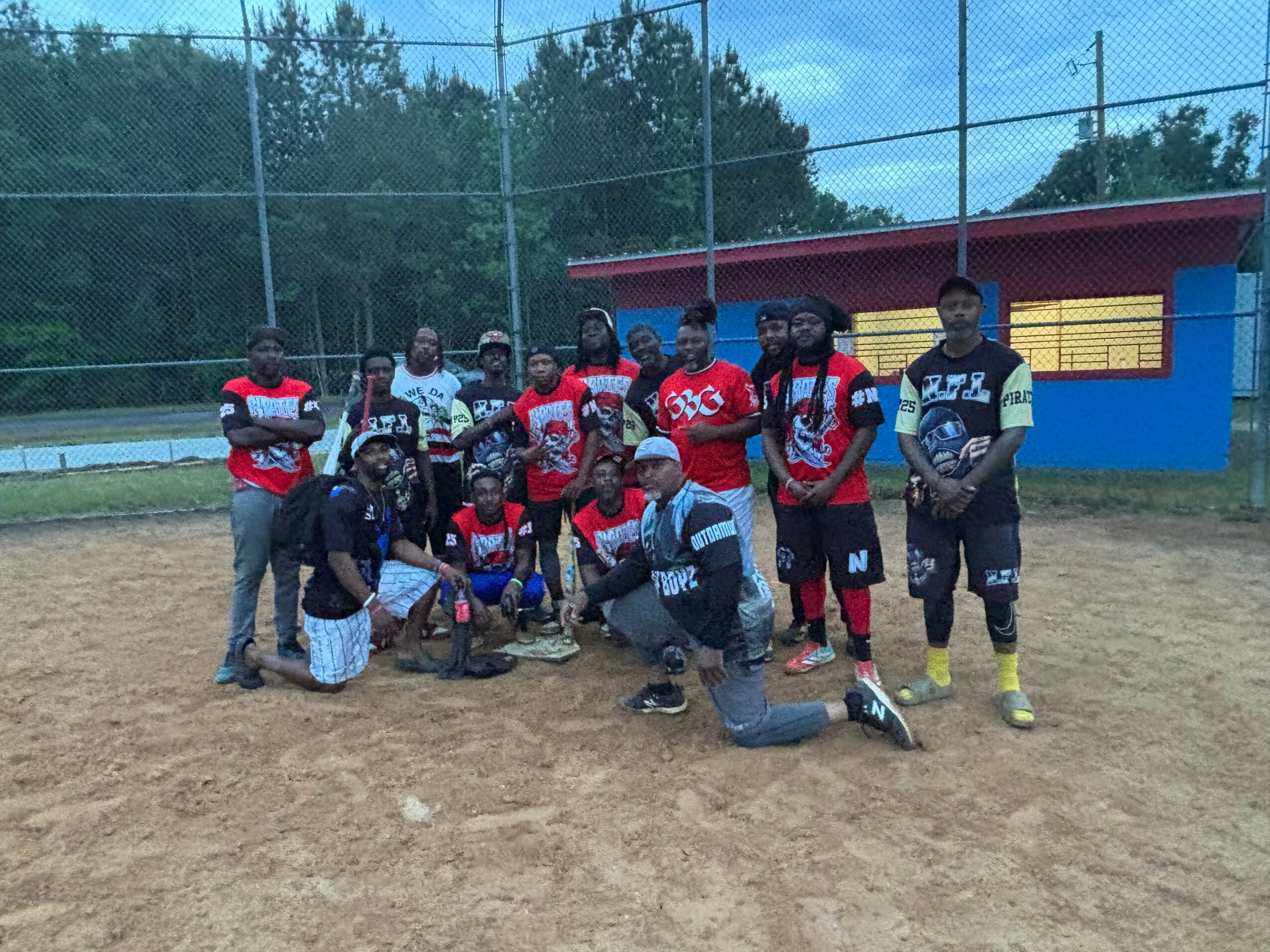 Group of baseball players and coaches on a field with a fence and colorful building in background.