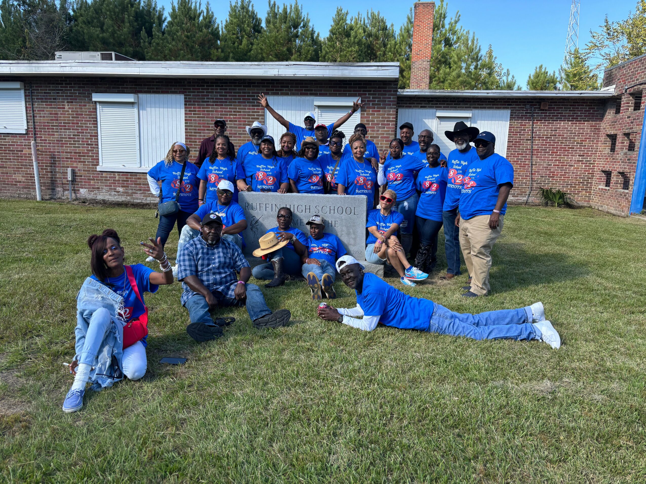 Group of people outdoors holding a banner, some kneeling, some standing, with a brick building and trees in background.