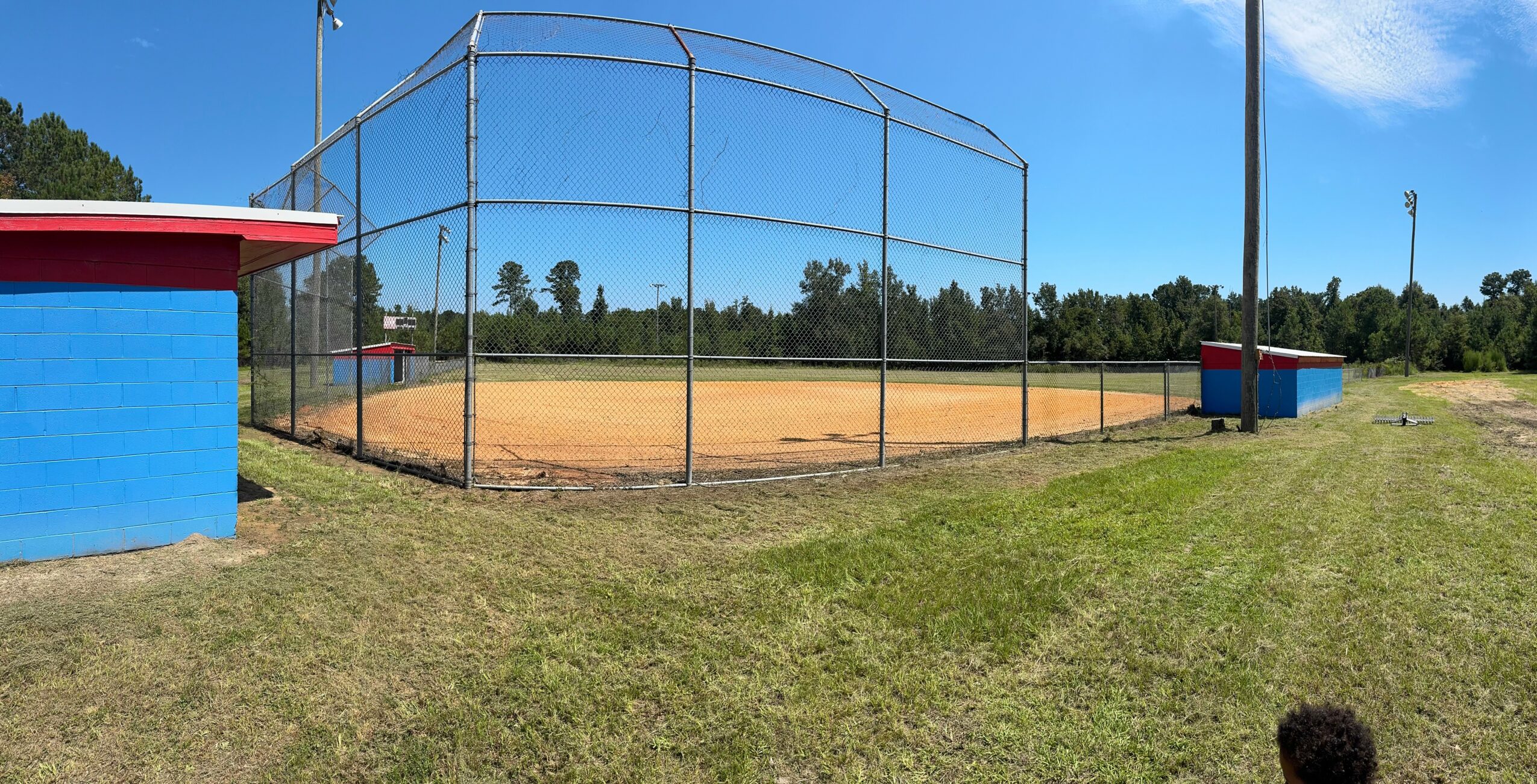 Baseball field with a chain-link fence, blue building, and grassy area under a blue sky.