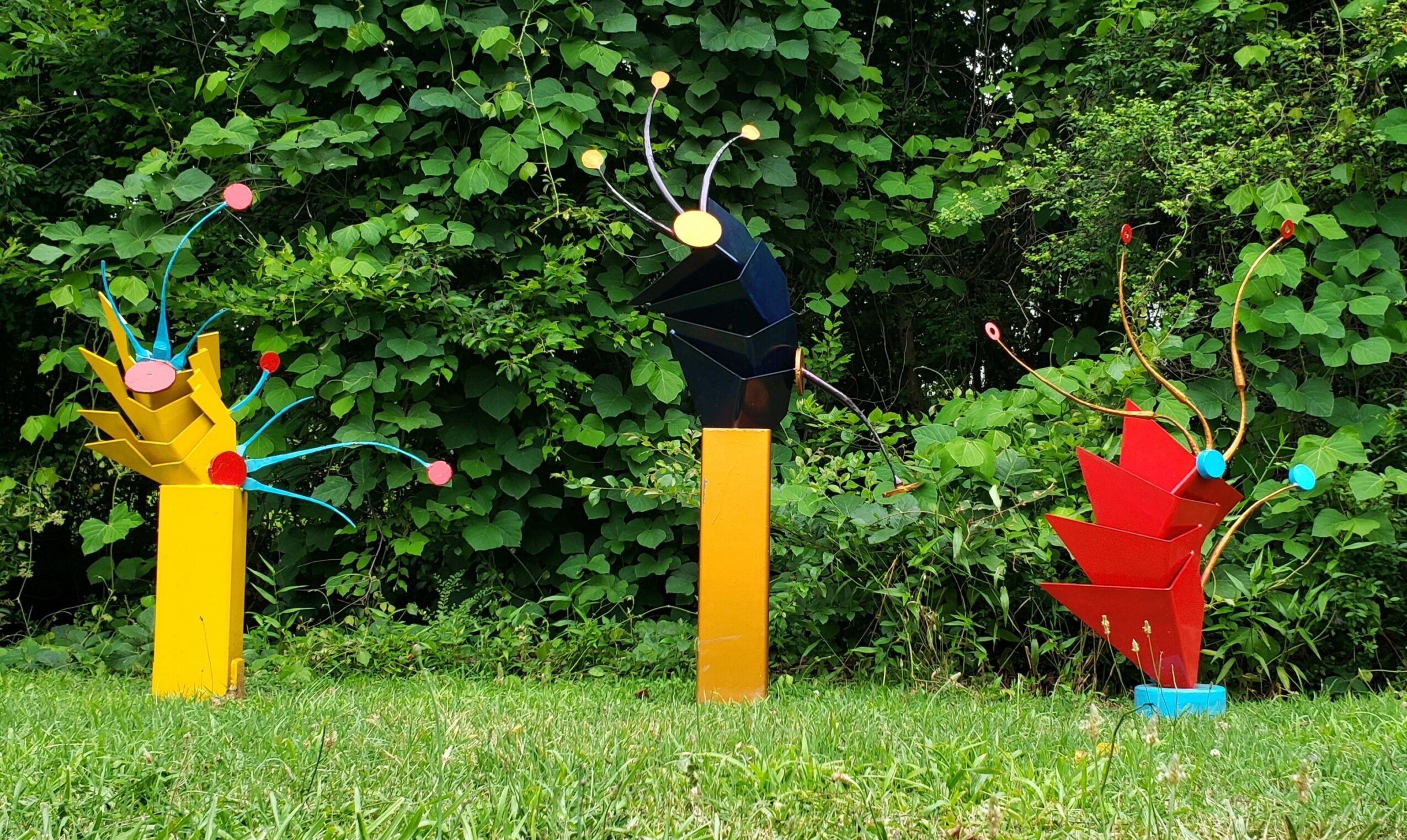 Three colorful insect sculptures with antennae and wings stand on grass in front of green bushes.