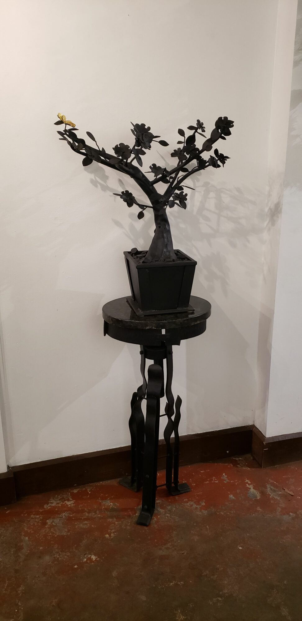 Sculpture of a tree with branches in a black rectangular planter, mounted on a stand, against a white wall.