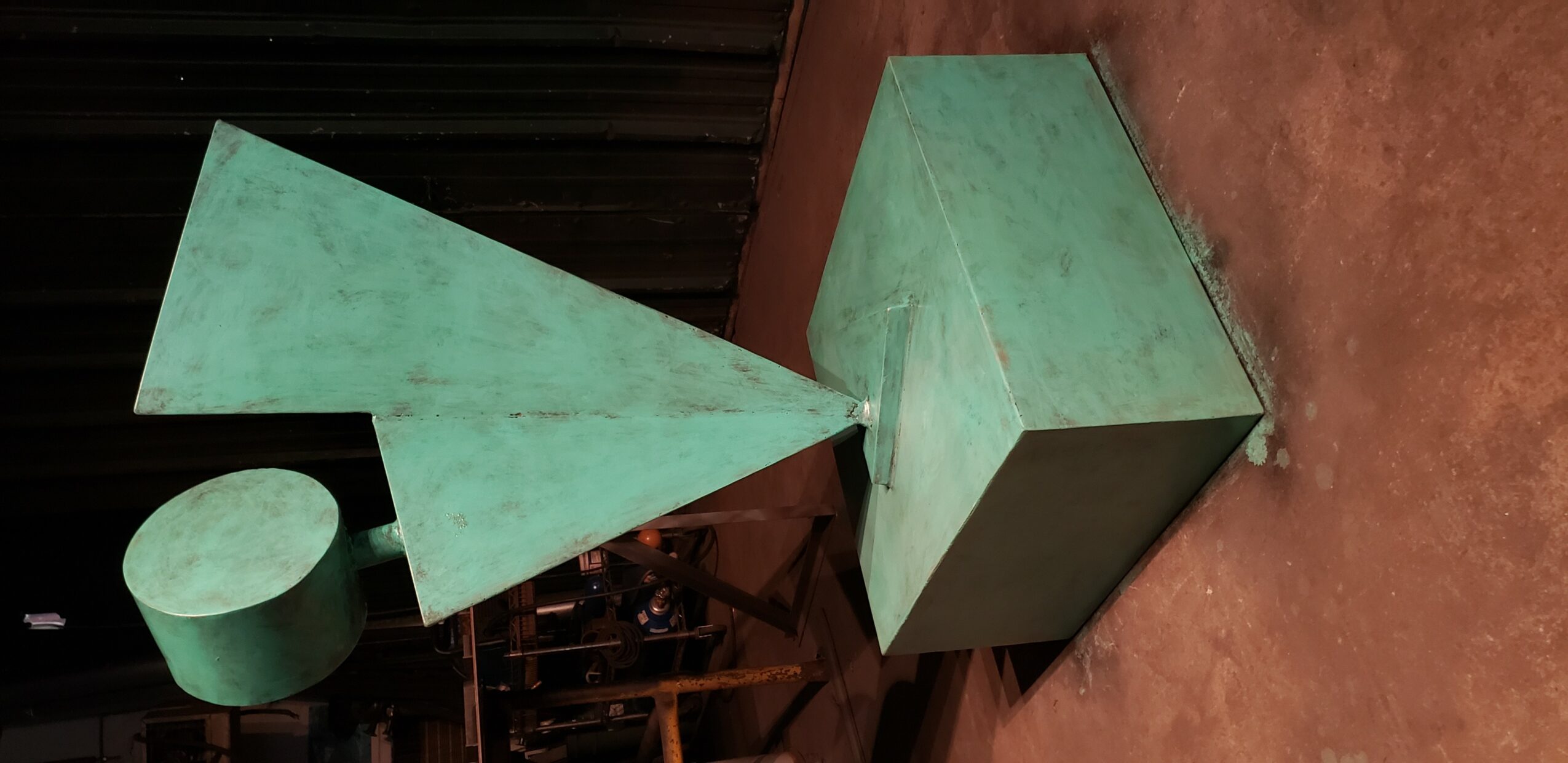 Green geometric sculpture with cone, triangle, and rectangular shapes on a brown floor.