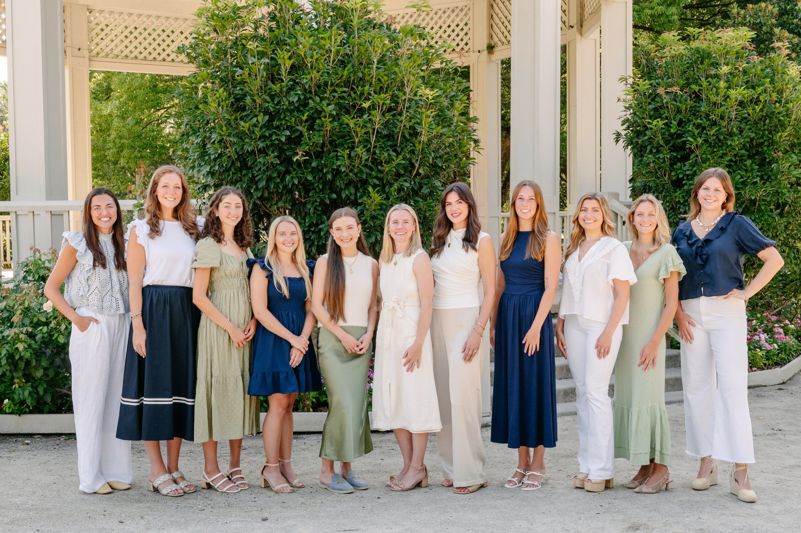 Group of eleven women standing outdoors in front of greenery and a white structure, smiling and dressed in casual summer clothing.