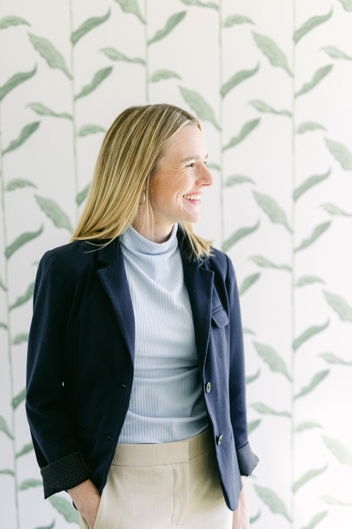 Woman with blonde hair smiling, wearing a navy blazer and light blue turtleneck, standing in front of a leafy patterned background.