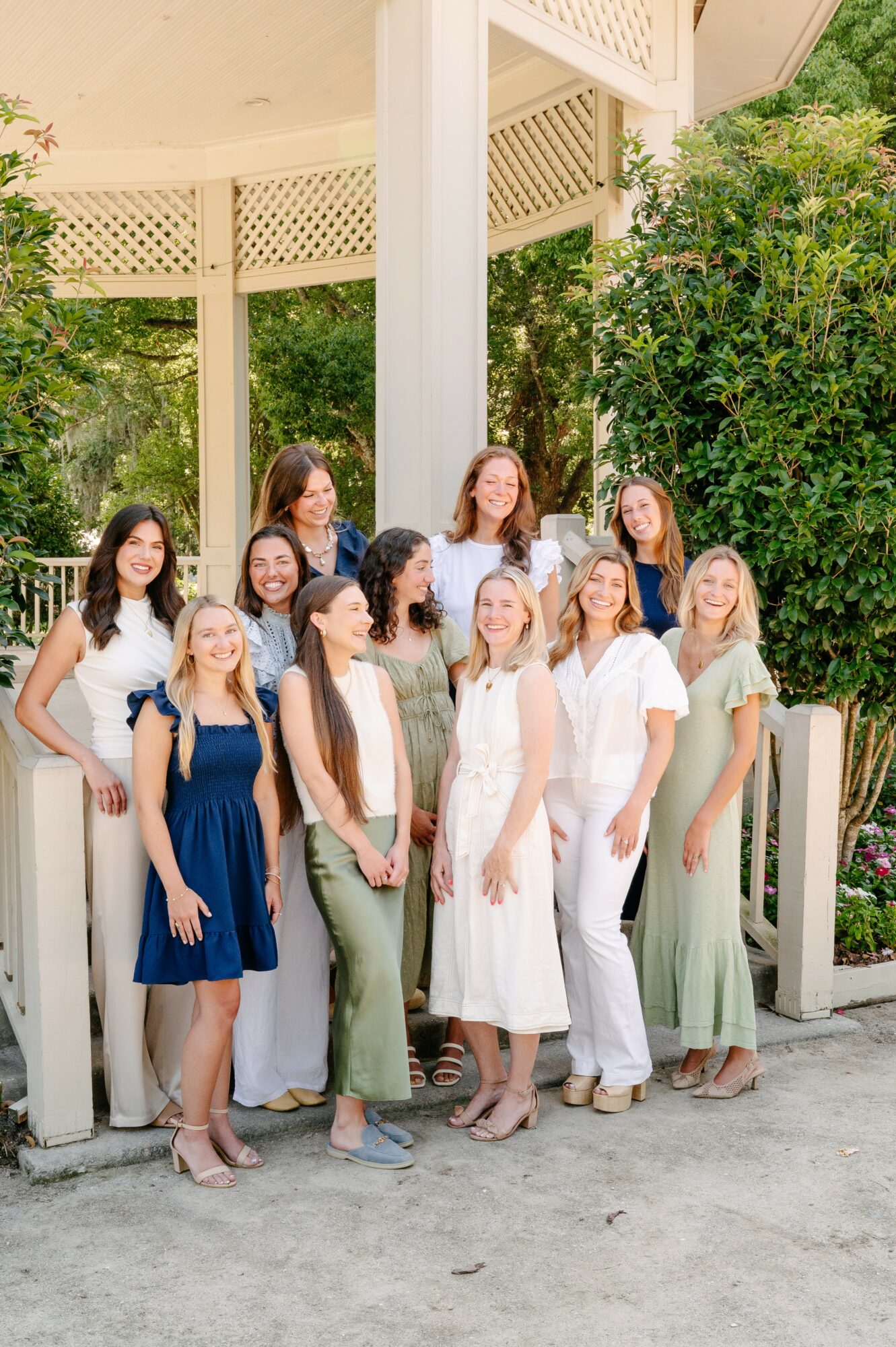 Group of ten women standing outdoors near a gazebo, smiling, with greenery in the background.