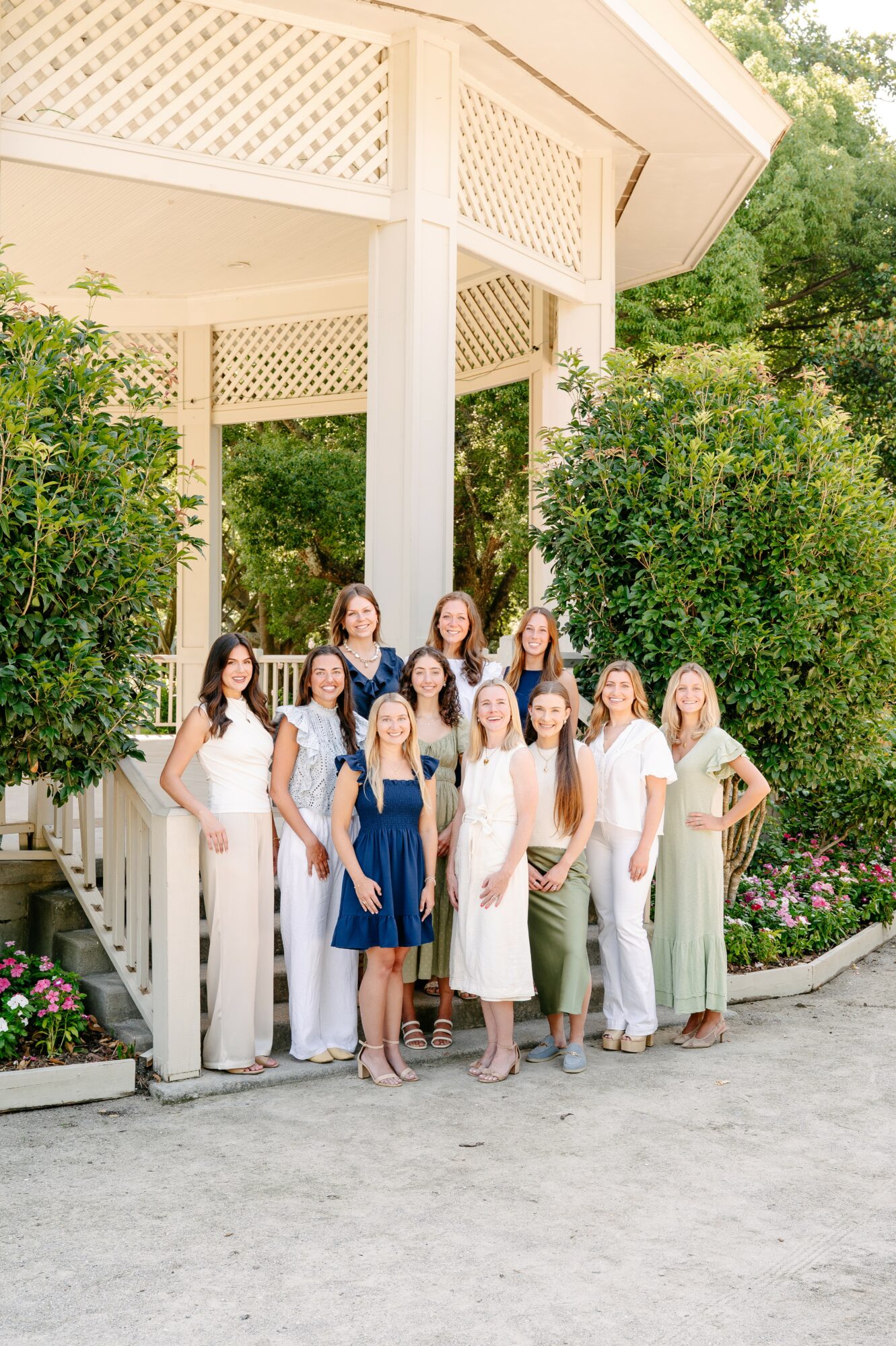 Group of women in white and blue dresses standing on a pathway near a gazebo with greenery.
