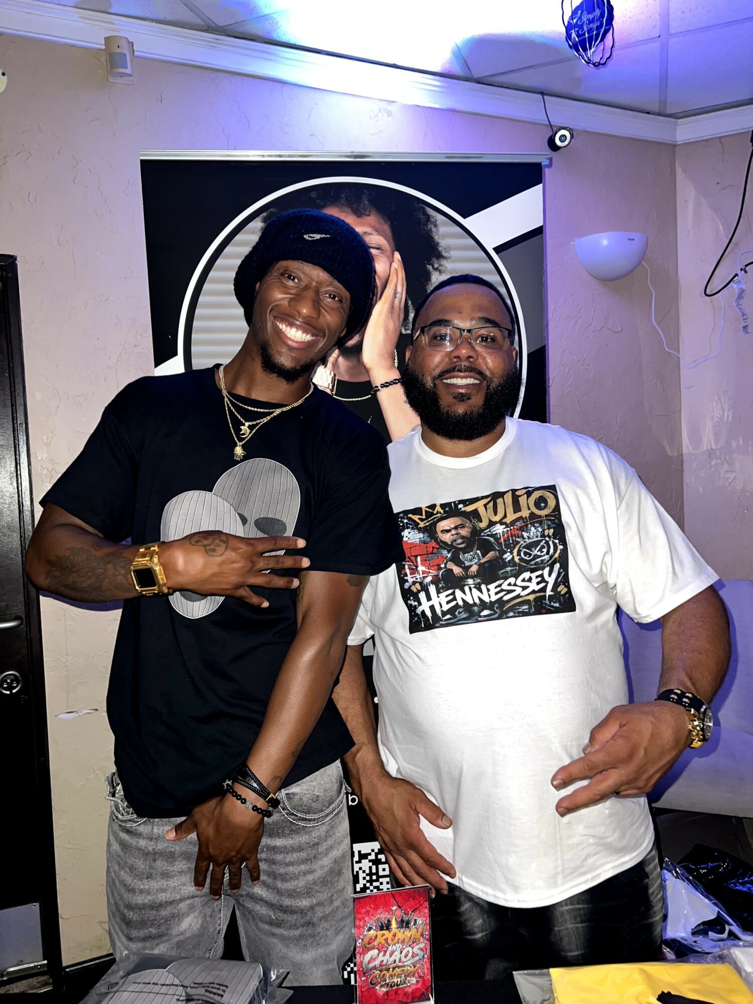 Two smiling men standing indoors, one wearing a black t-shirt and the other a white t-shirt with a graphic, in front of a screen.