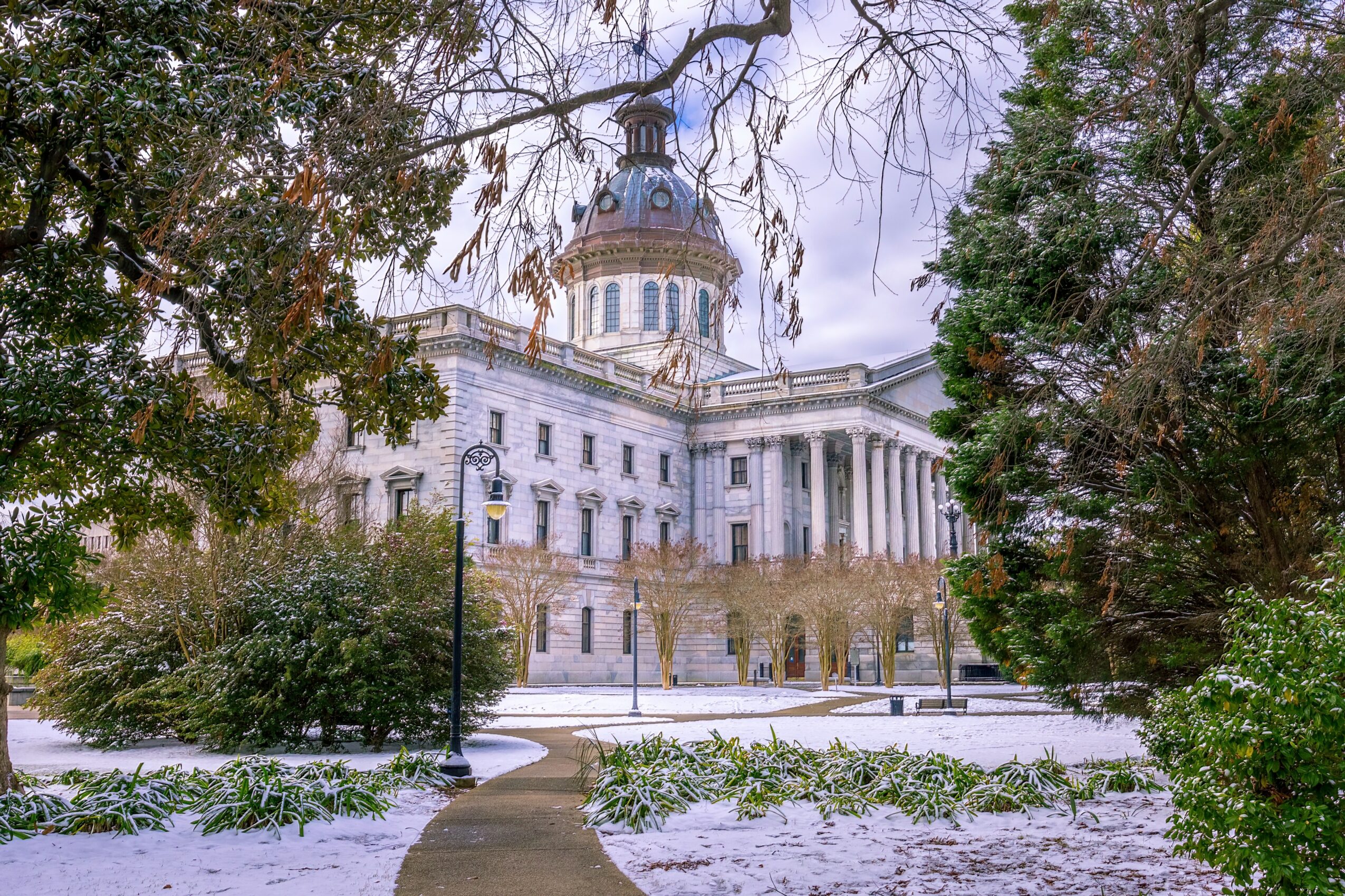 A large historic building with a dome, surrounded by trees and a pathway, under a cloudy sky.