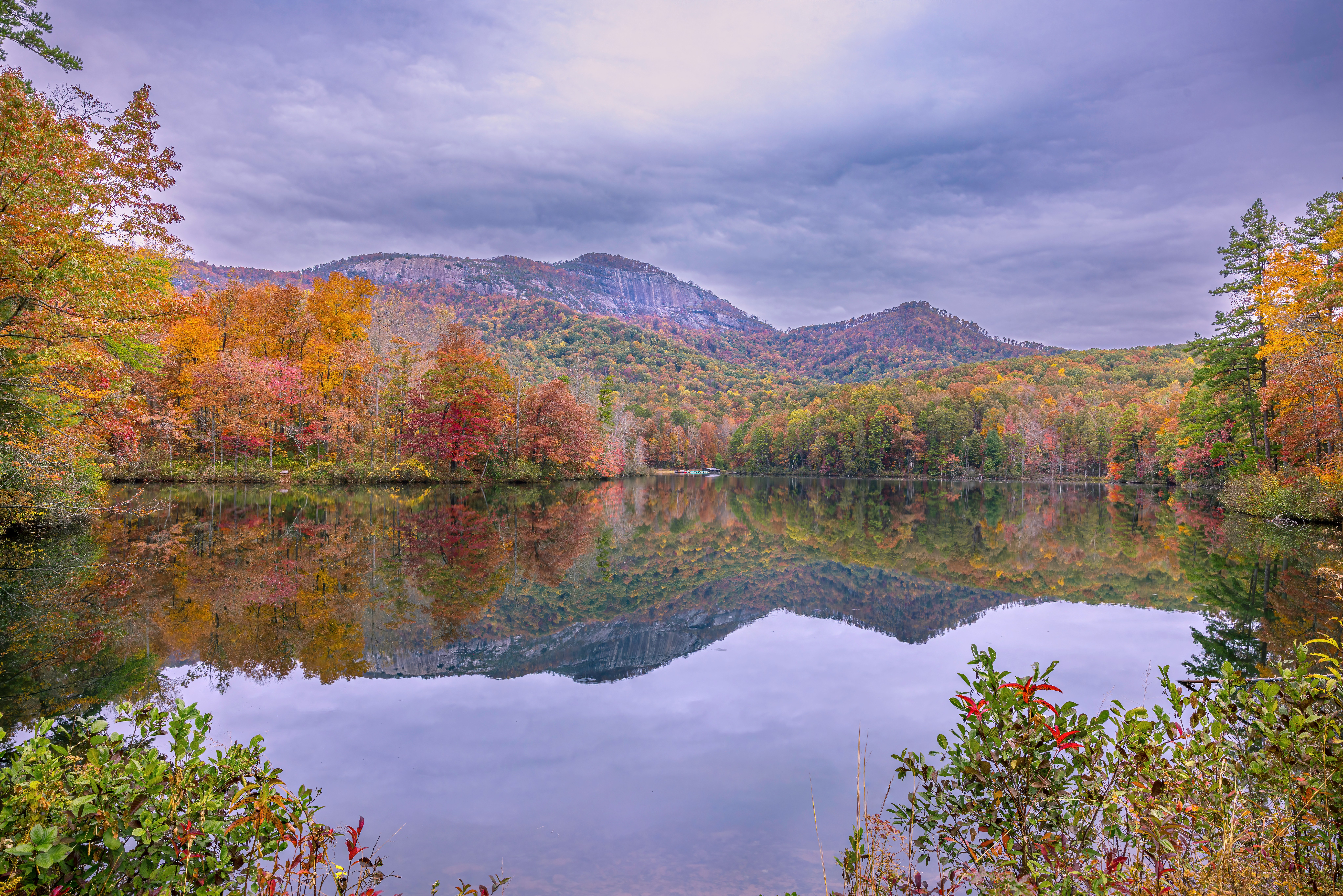 Scenic view of a lake surrounded by colorful autumn trees and mountains in the background, with a cloudy sky.