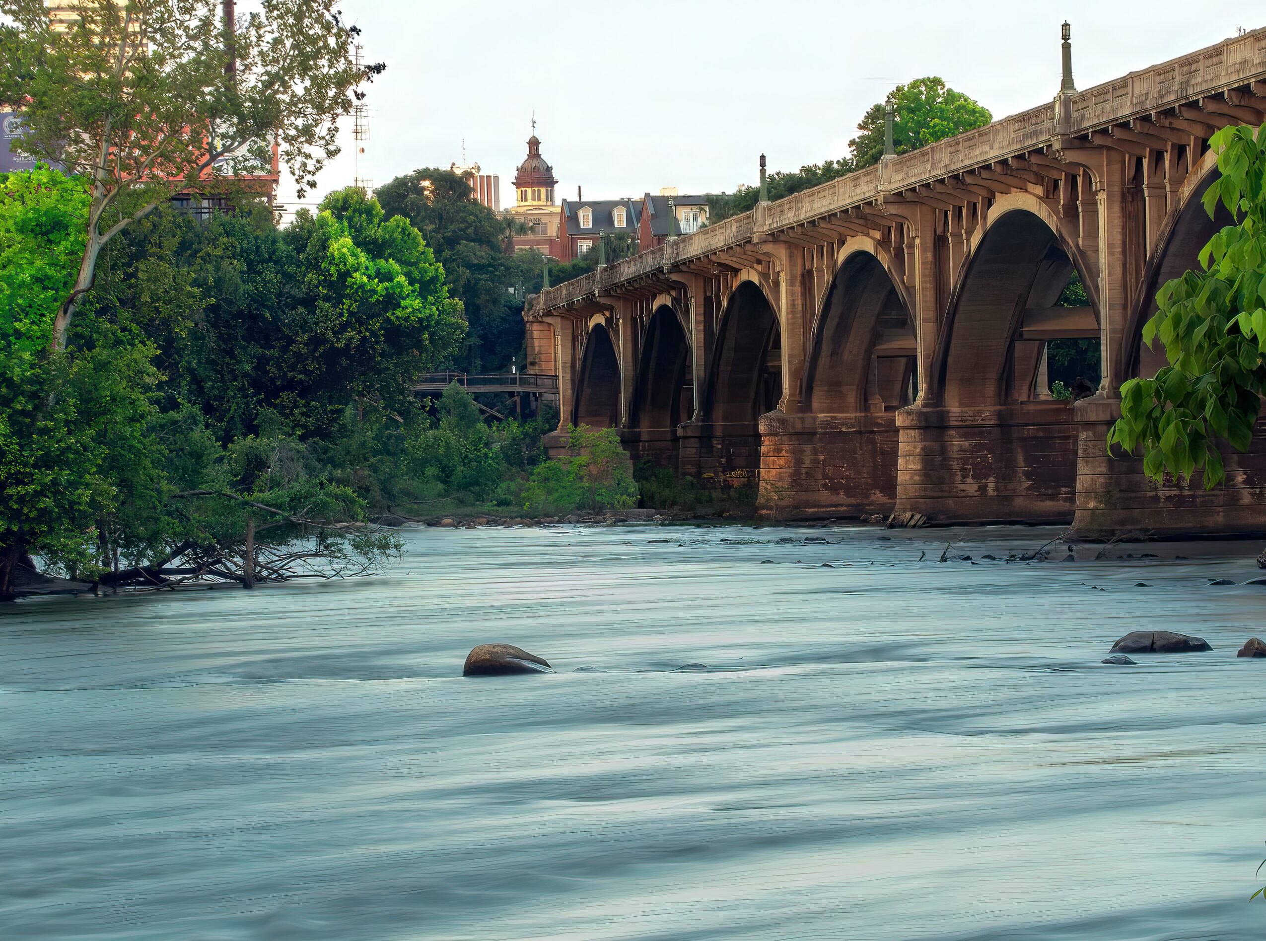 Bridge over a river with trees and buildings in the background, water flowing smoothly in the foreground.