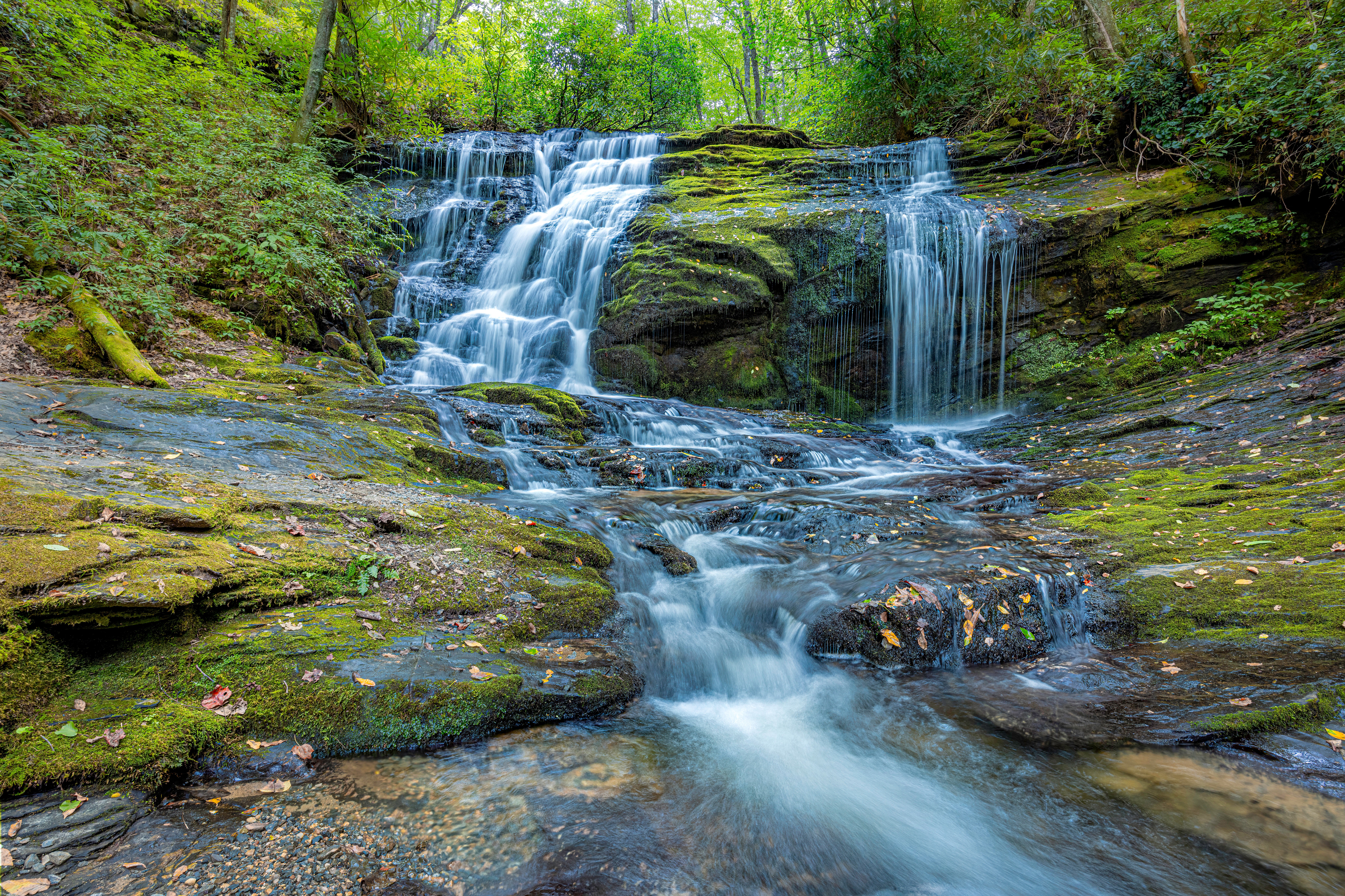 Waterfall flowing over rocks in a lush green forest, with water cascading down multiple levels and trees in the background.