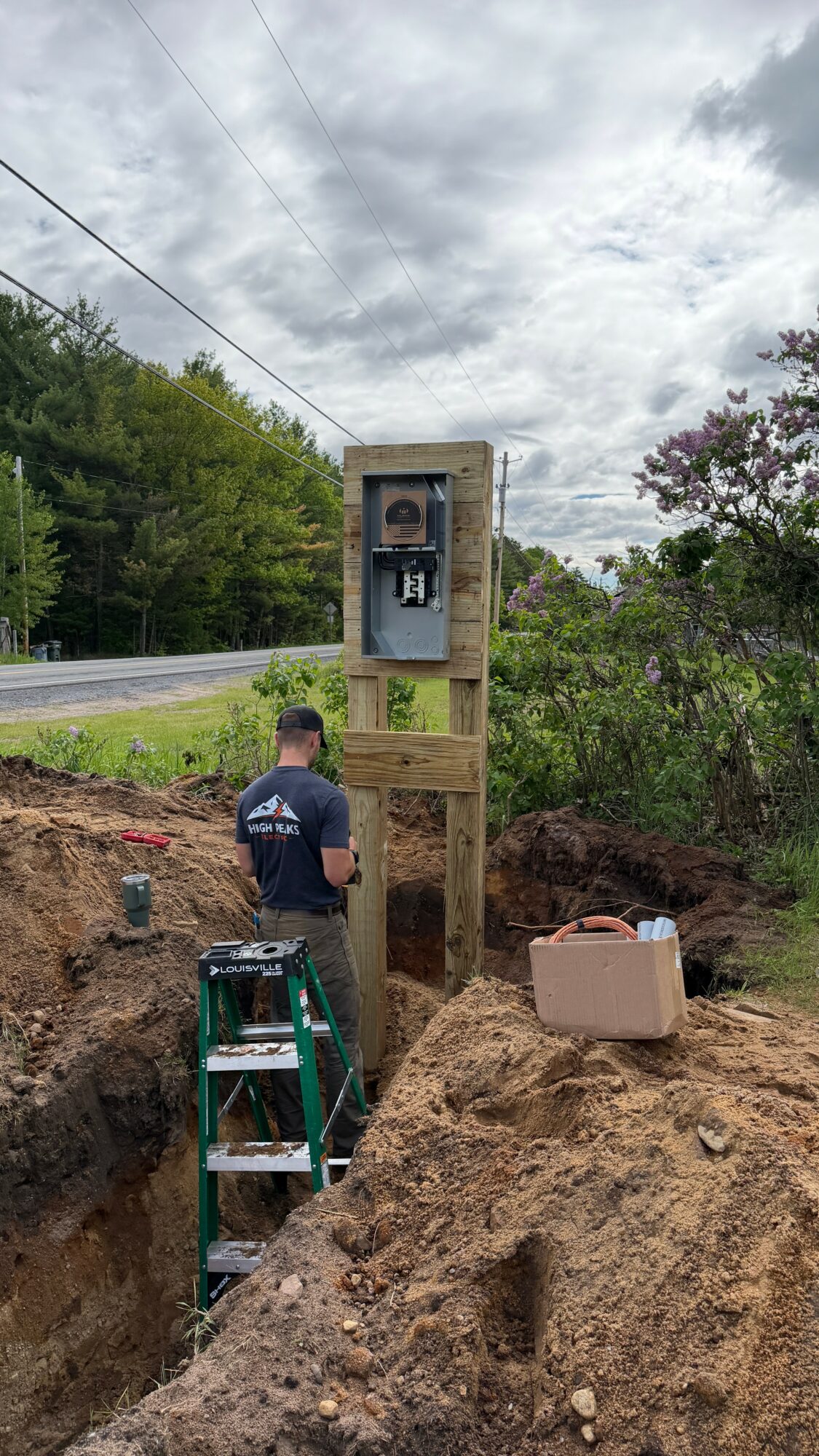 Person working on electrical box outdoors, ladder nearby, dirt and trees around, cloudy sky above.