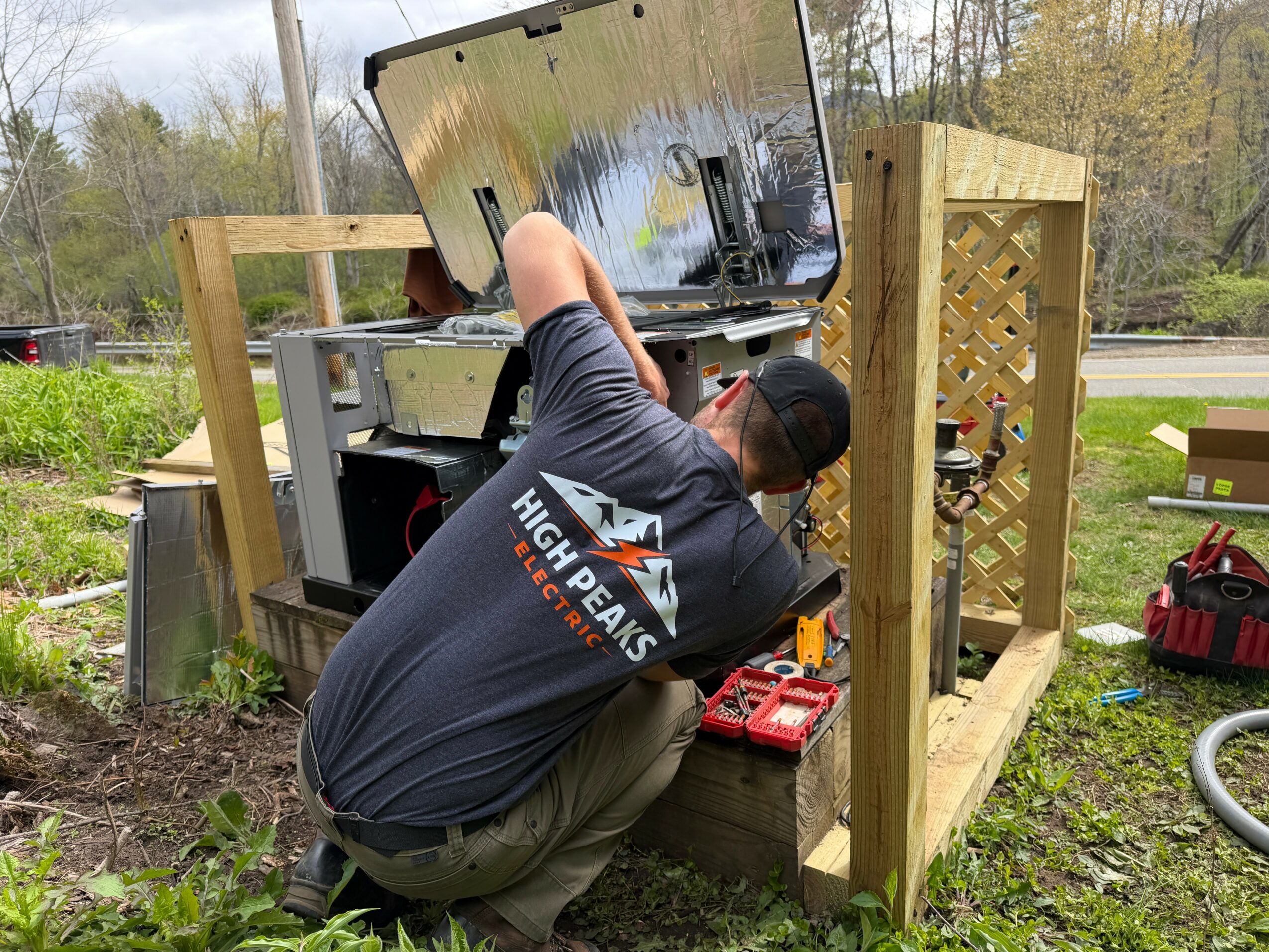 Person working on a computer outdoors, surrounded by wooden structures and tools, with a garden and trees in the background.