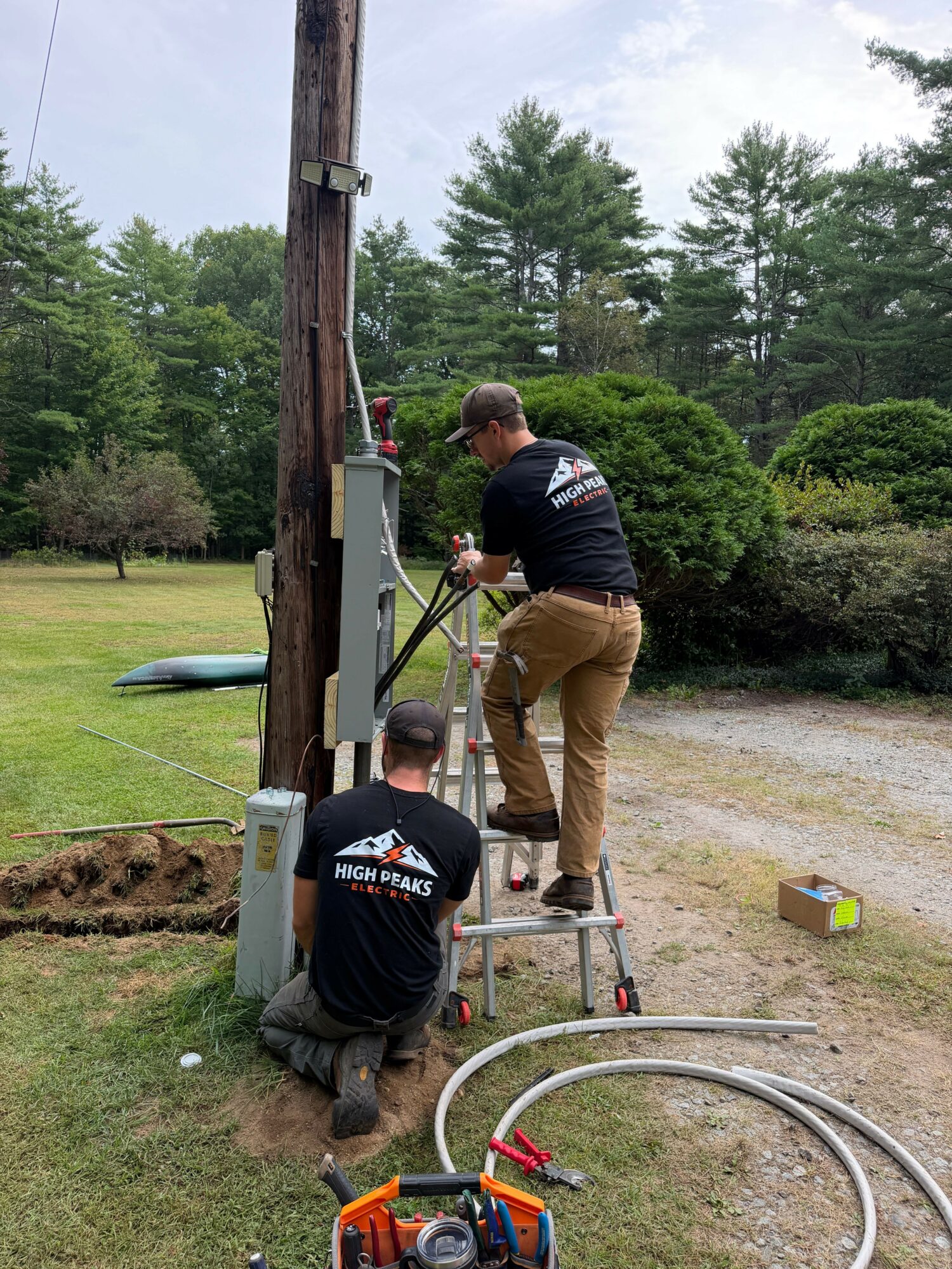 Two workers installing or repairing equipment outdoors near a tree and bushes, with tools and cables on the ground.