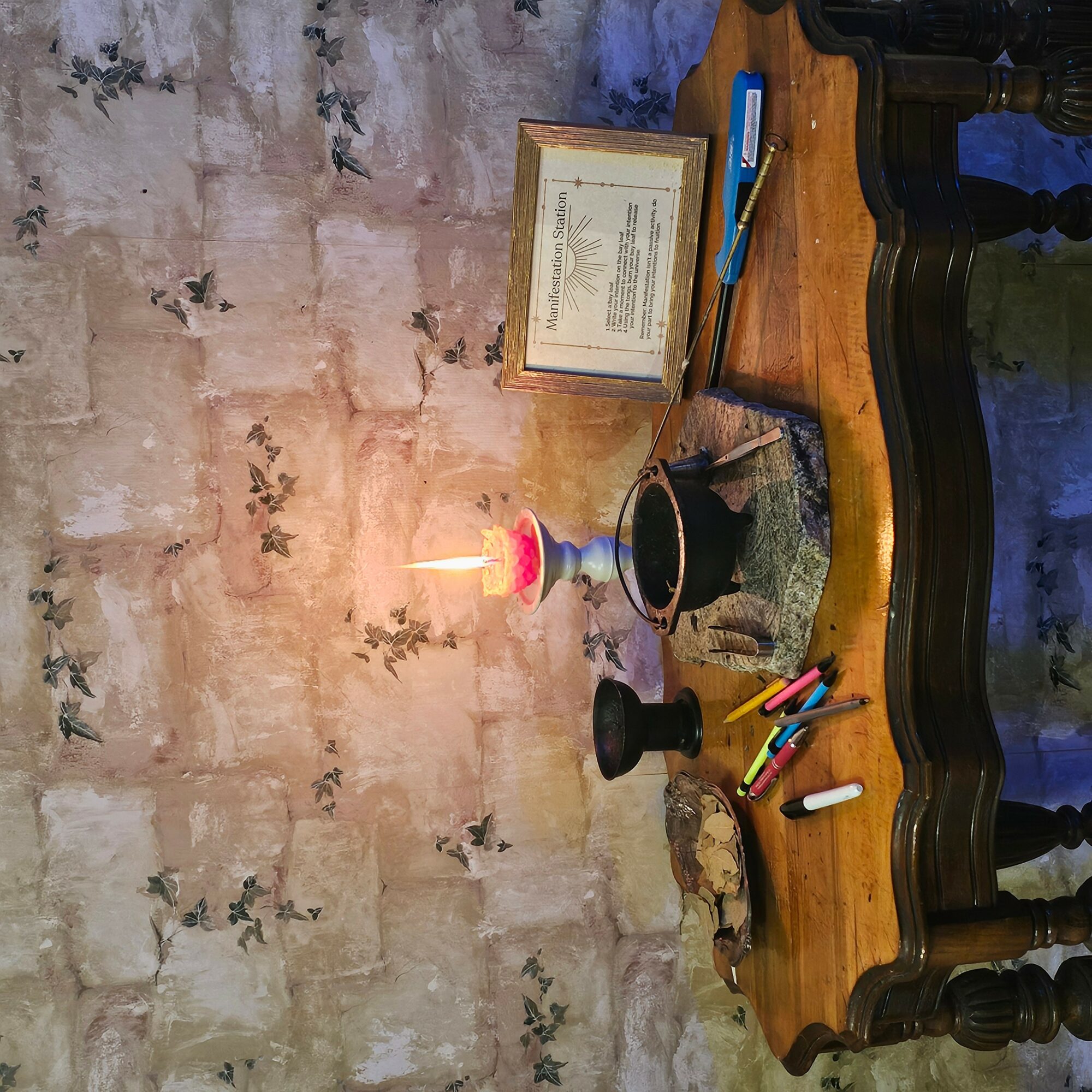 Table with a lit candle, framed document, and colorful markers on a textured wall background.