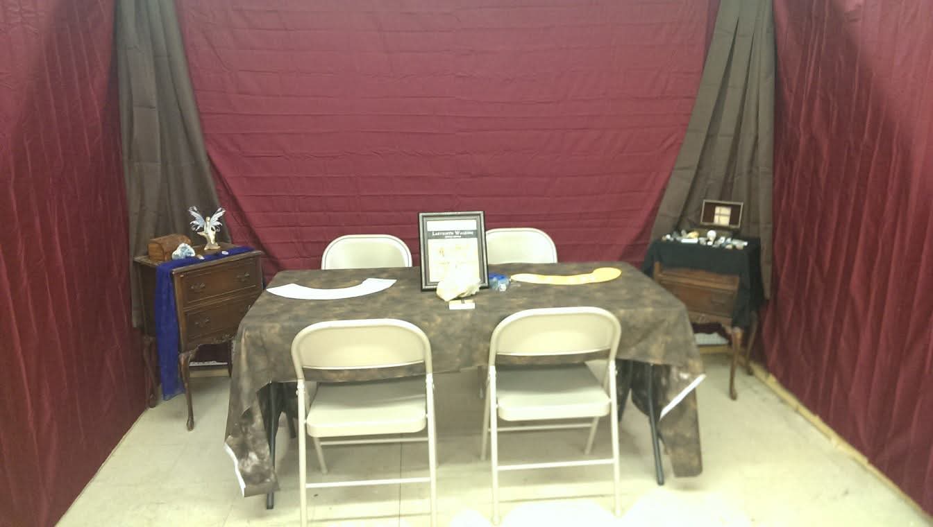 Rectangular table with six chairs, a framed document, and decorative items, set against red curtains with black drapes.