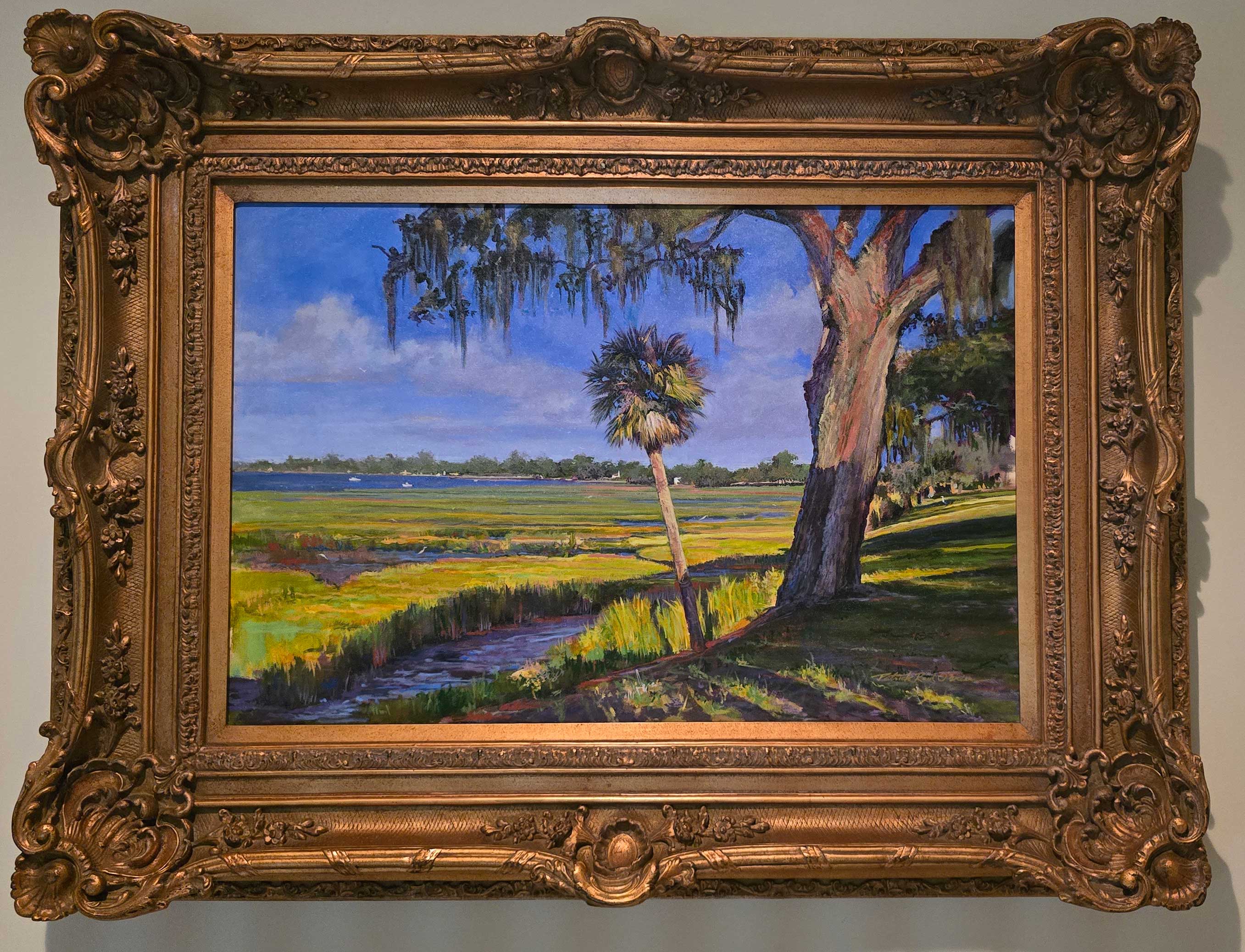 Landscape painting of a marsh with a large tree, a smaller palm, and a blue sky with clouds, framed in ornate gold.