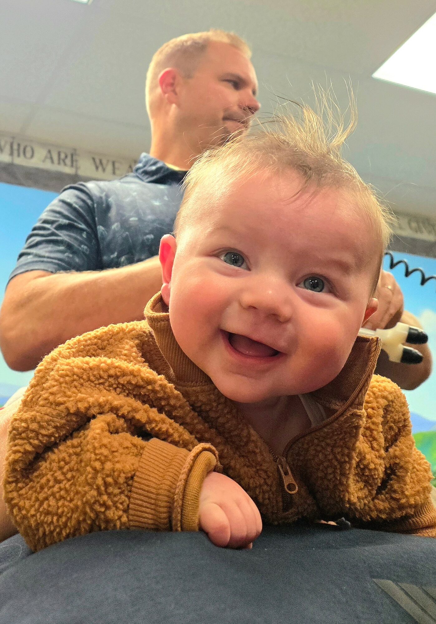 Smiling baby in a brown fuzzy jacket with a person in the background, indoors.