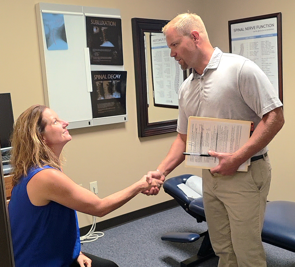 A woman sitting and shaking hands with a man holding a folder in a medical office. Two framed posters are on the wall.