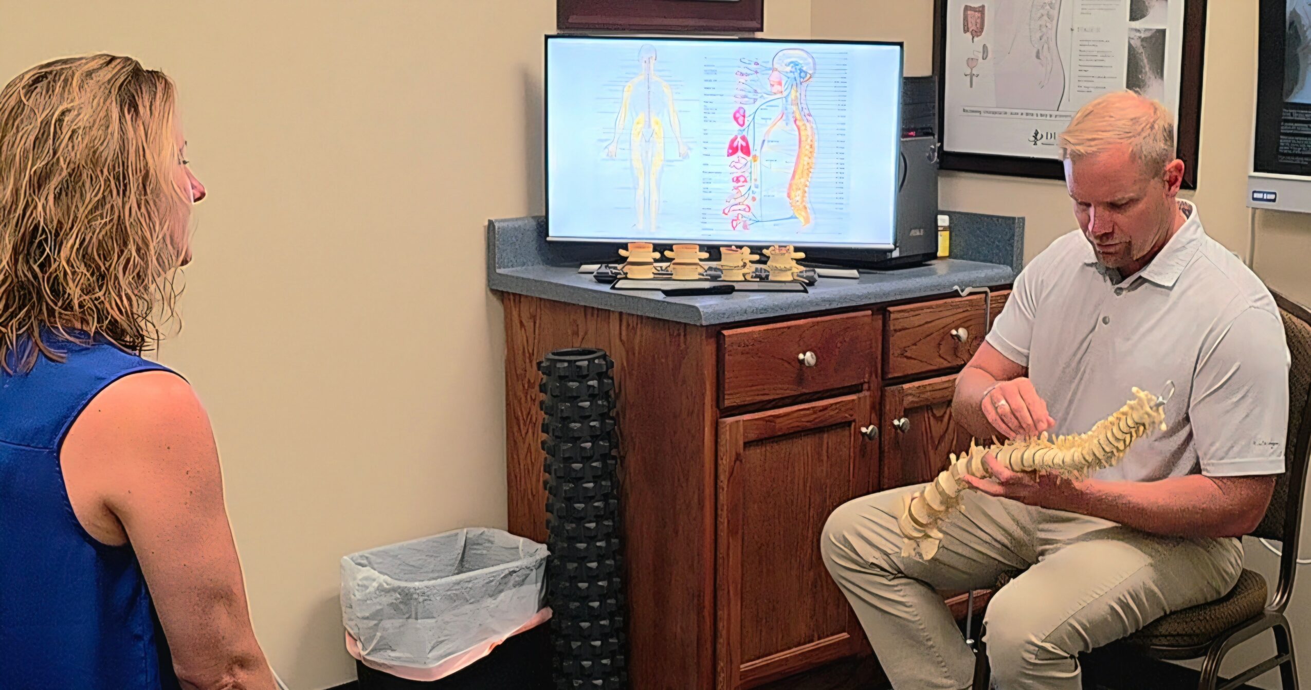 A man holds a model of a spine while a woman watches in a room with a television and cabinets.