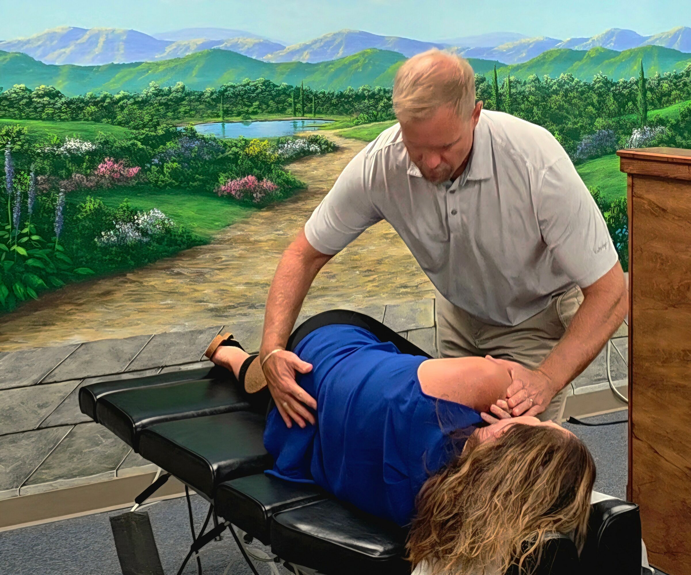 A man performs a chiropractic adjustment on a person lying face down on a treatment table, with a painted landscape background.
