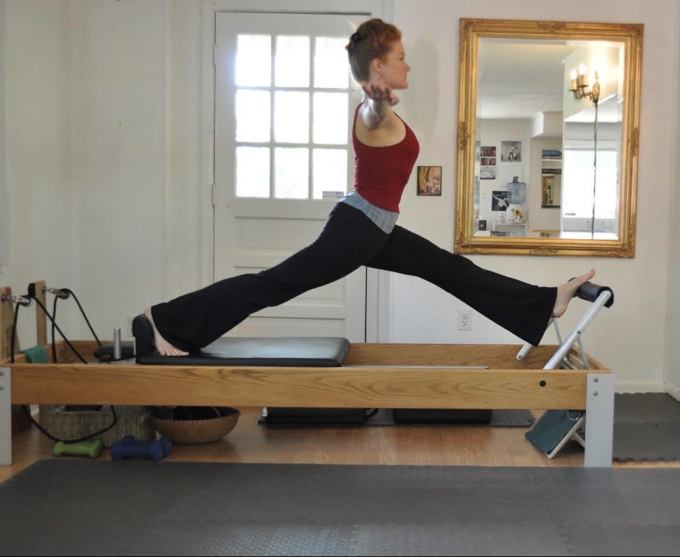 Woman in red top and black pants stretching on a wooden platform in a room with a window, mirror, and framed pictures.