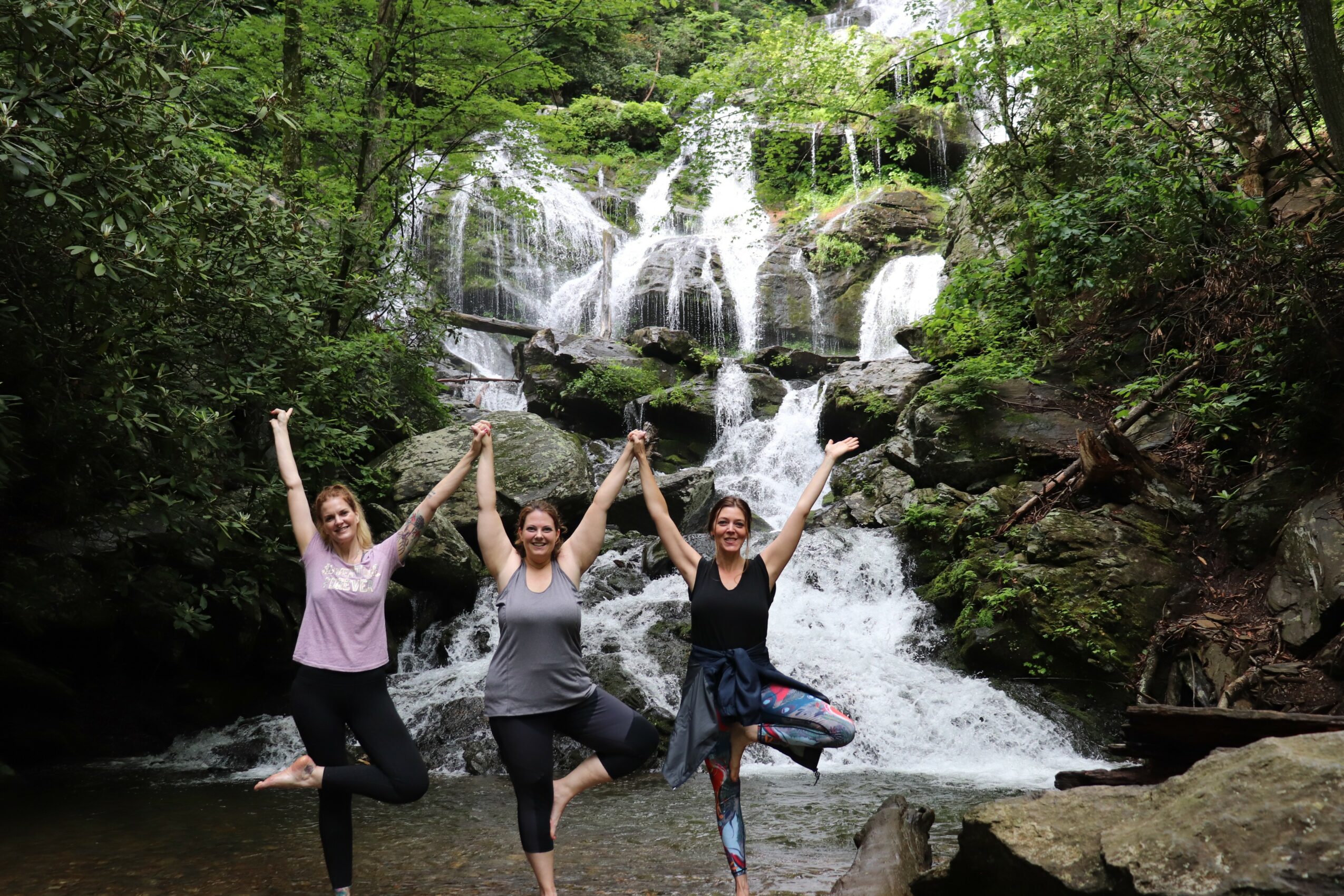 Three women standing on rocks in front of a waterfall, holding hands and raising arms, surrounded by trees.
