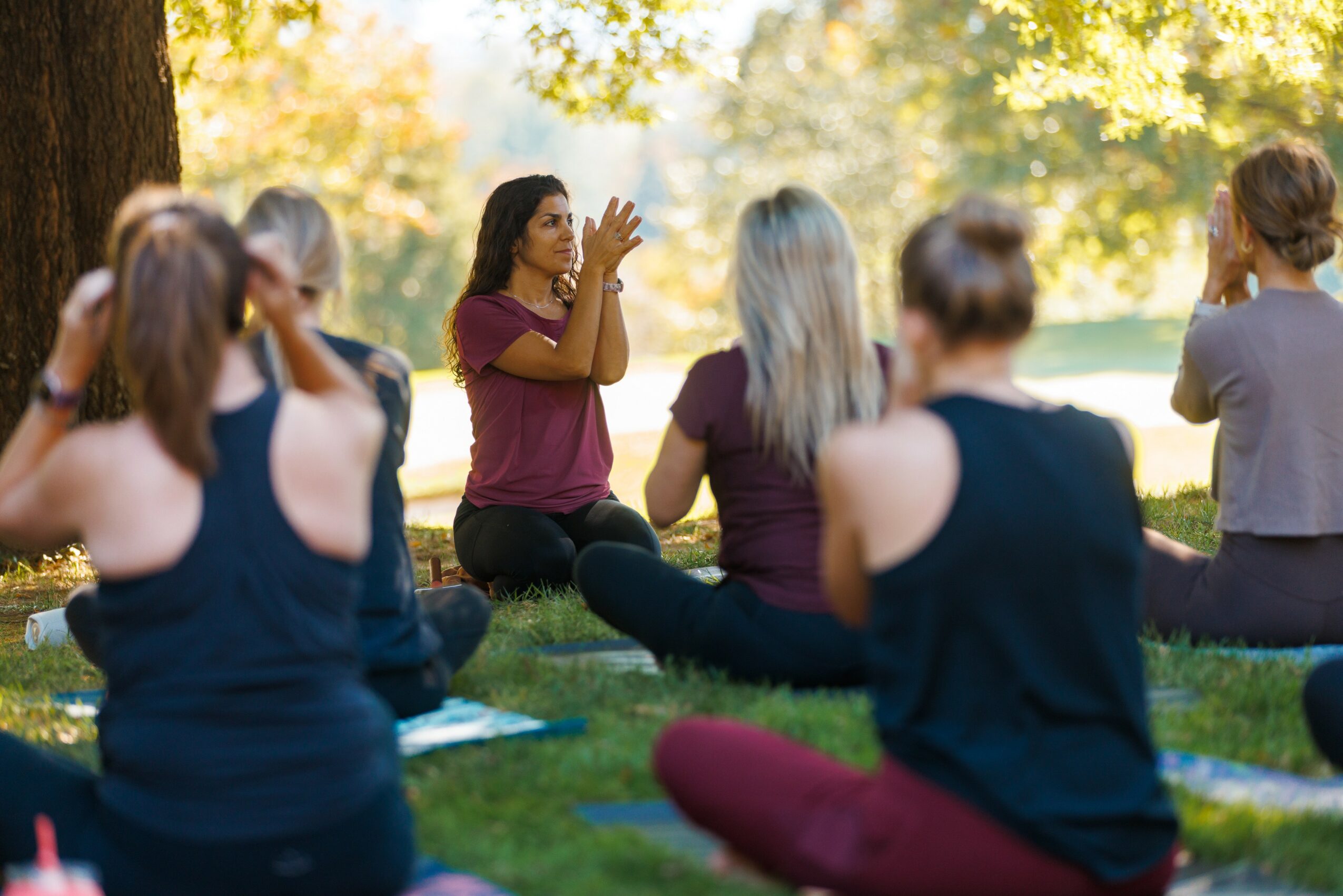 Group of people sitting outdoors on grass, participating in a meditation or yoga session, with trees and sunlight in background.