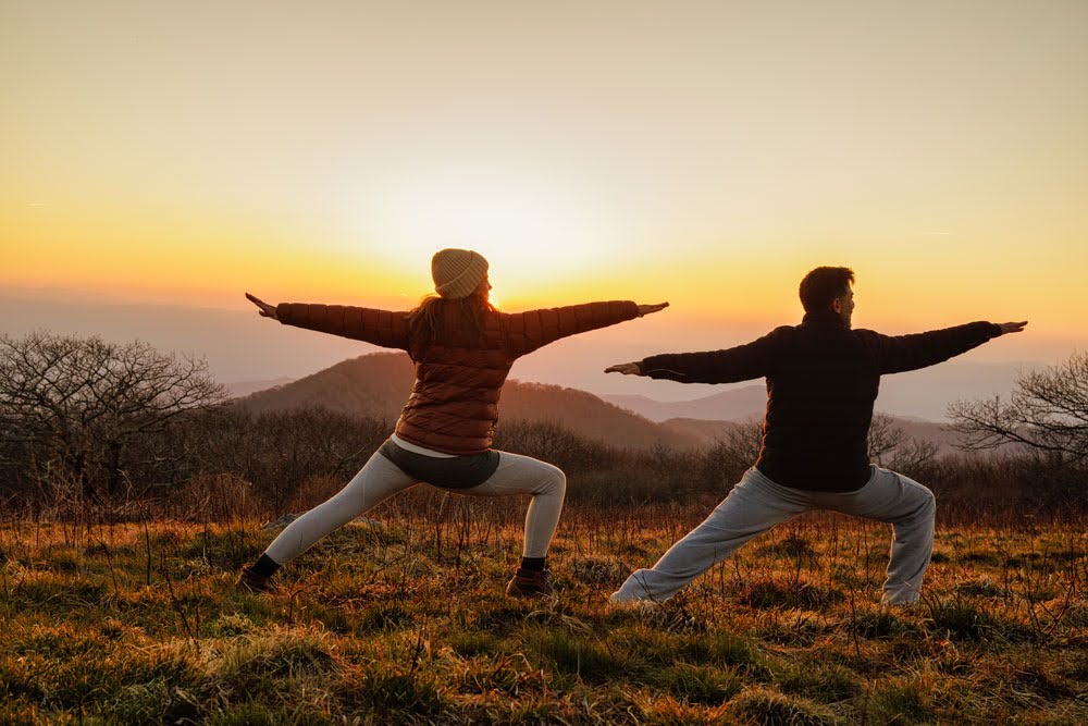 Two people practicing yoga outdoors at sunset, standing in warrior pose with arms extended, in a grassy field with trees and mountains.