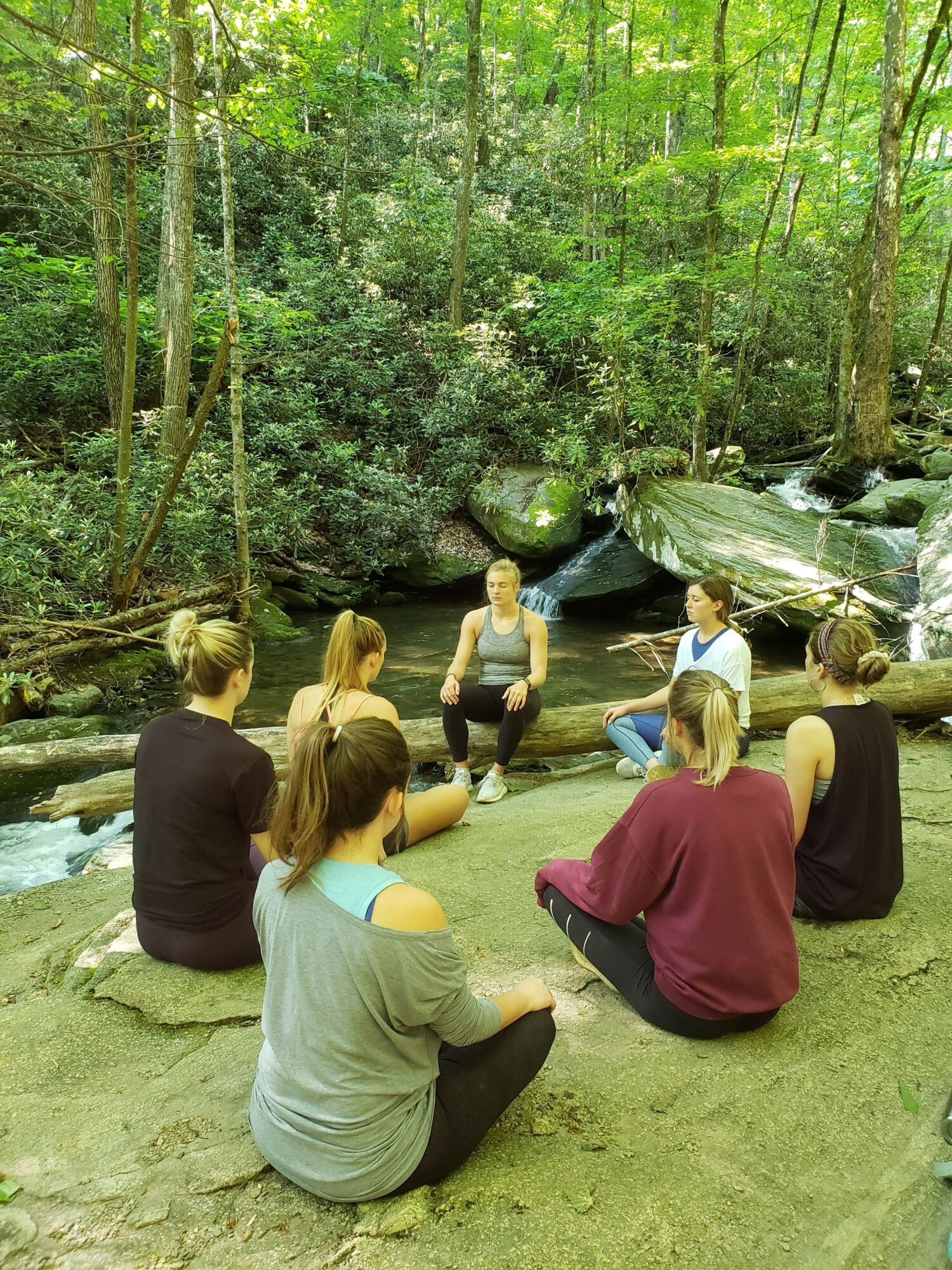 Group of people sitting on a rock in a forest near a stream, surrounded by trees and greenery.