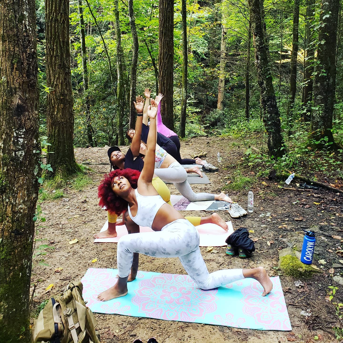 Four people practicing yoga outdoors in a forest, performing various poses on mats, with trees and greenery around.