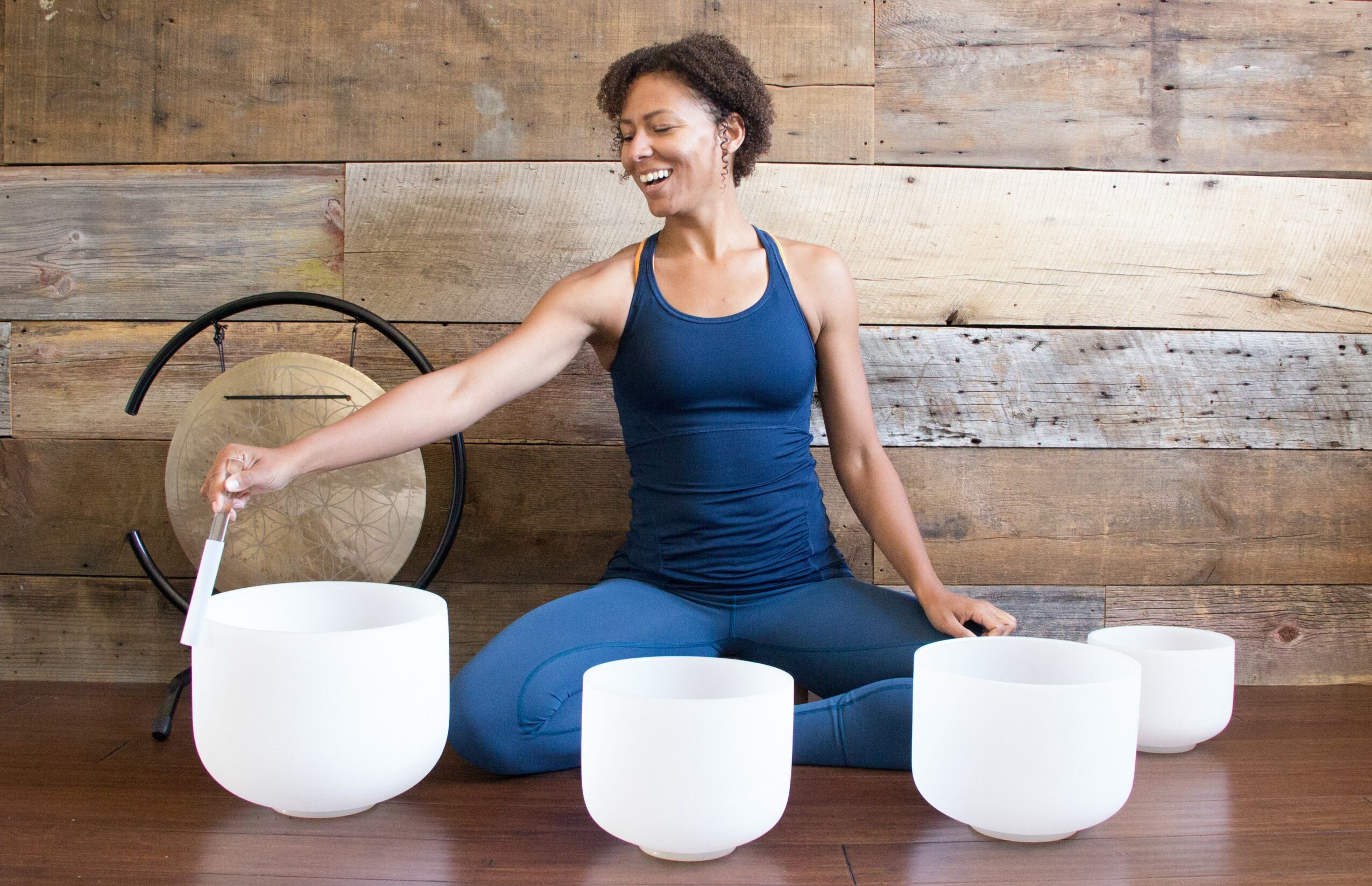 Woman sitting cross-legged on wooden floor, smiling, playing crystal singing bowls, with wooden wall background.