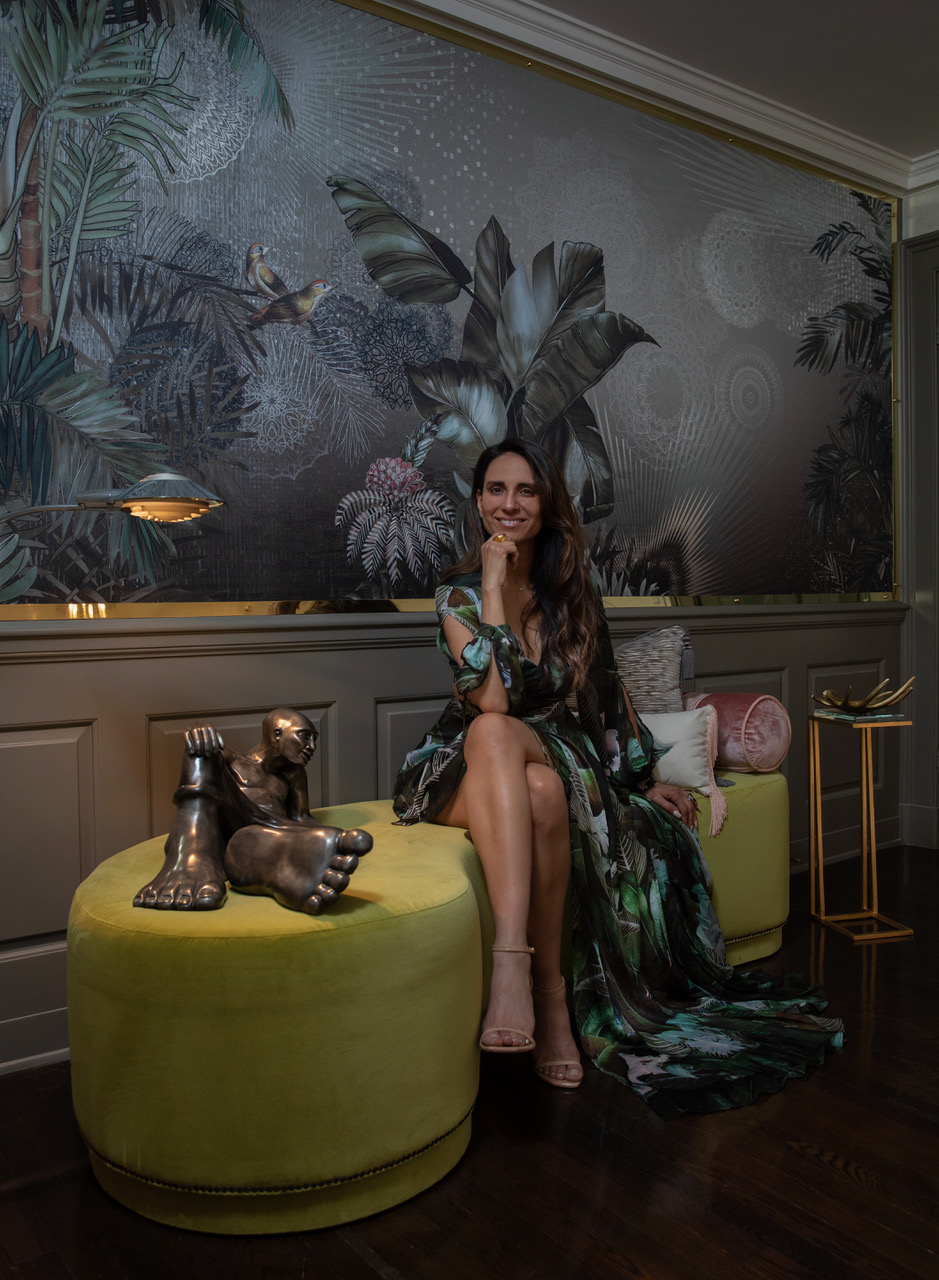 Woman sitting on a yellow ottoman with decorative dog sculptures, in a room with tropical wallpaper and dark wooden floor.