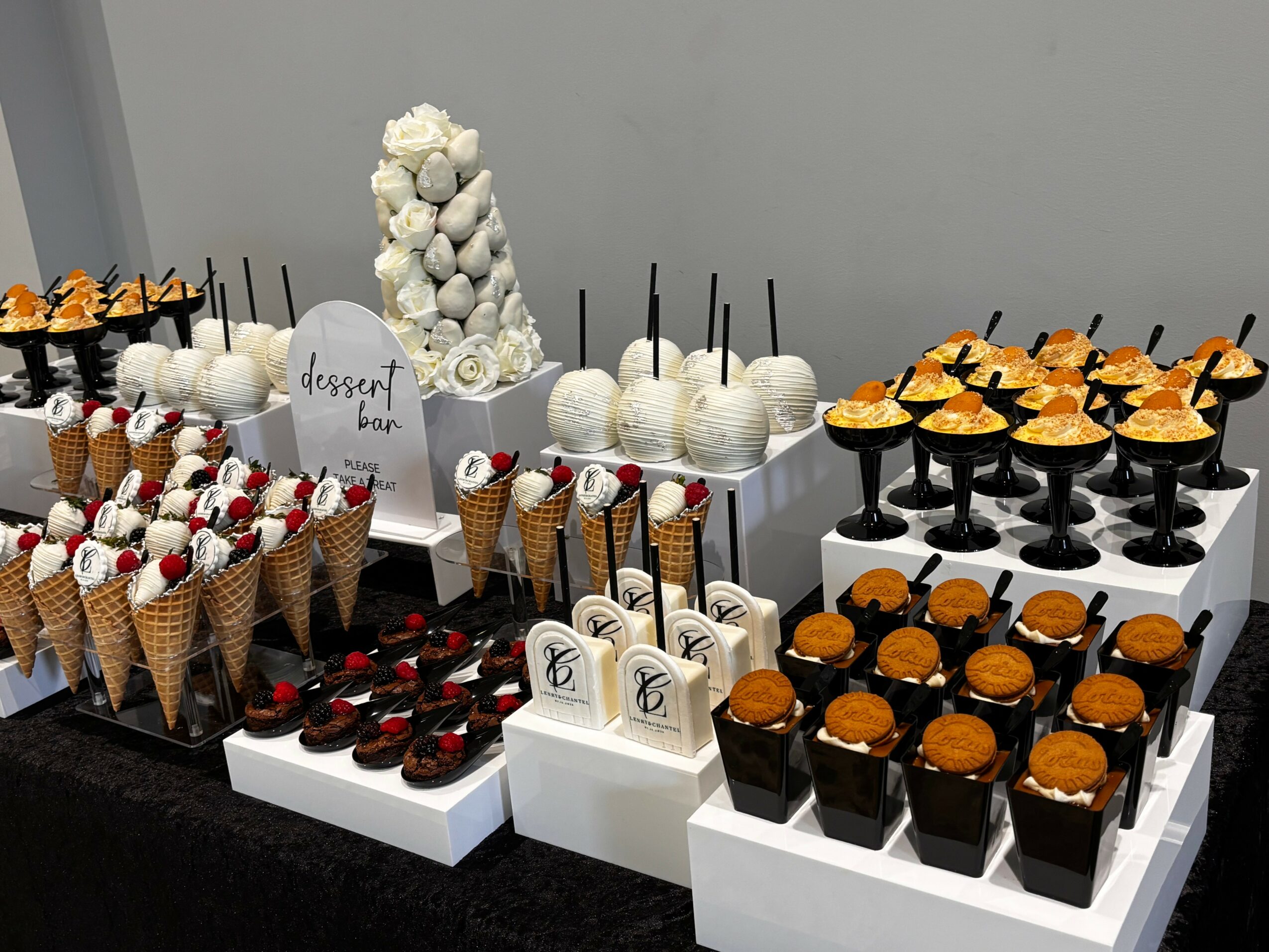Assorted desserts including cake pops, cupcakes, and chocolates arranged on white display stands with a white flower decoration in the background.