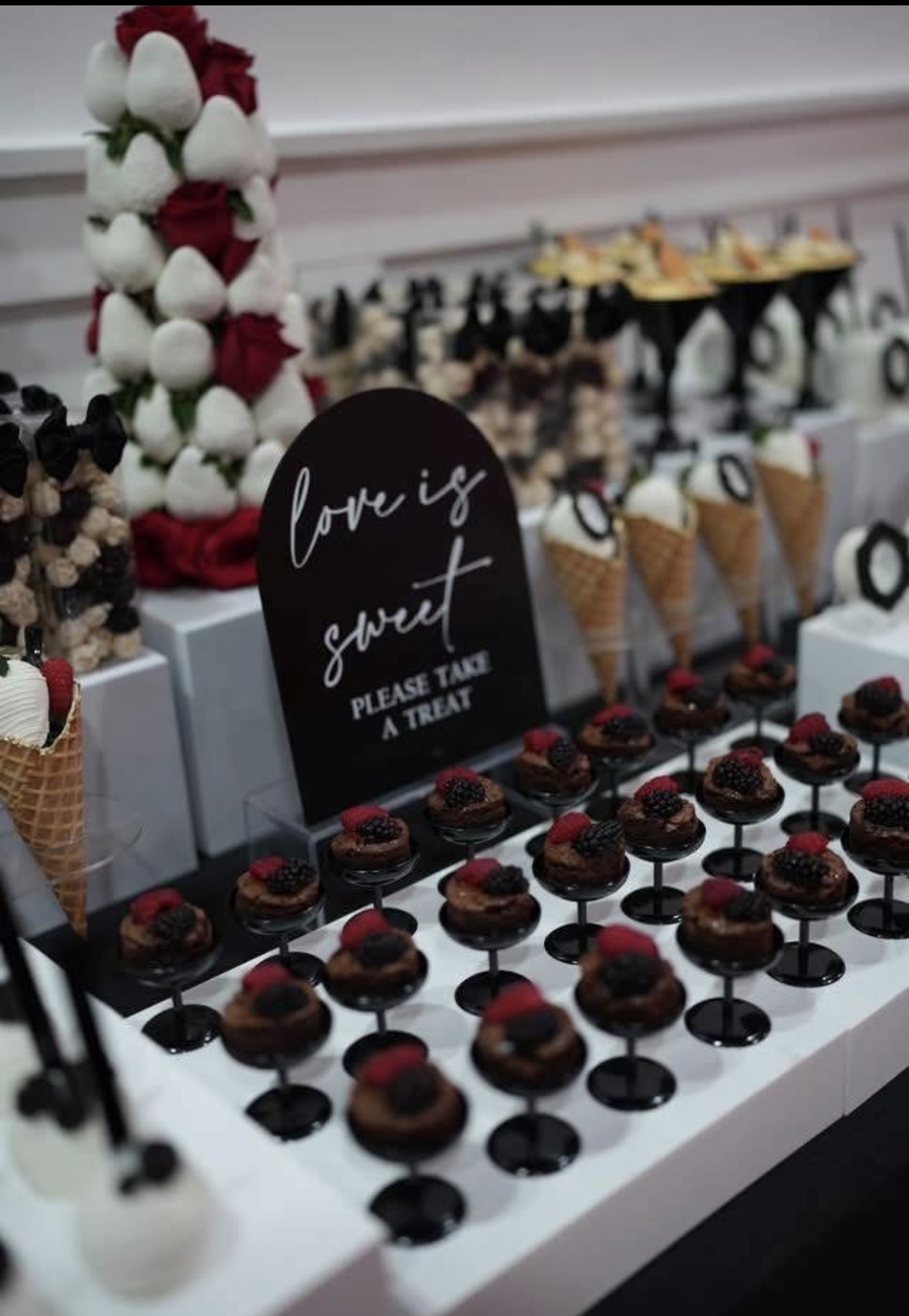 Dessert table with chocolates, ice cream cones, and a sign reading 'love is sweet, please take a treat,' decorated with flowers.