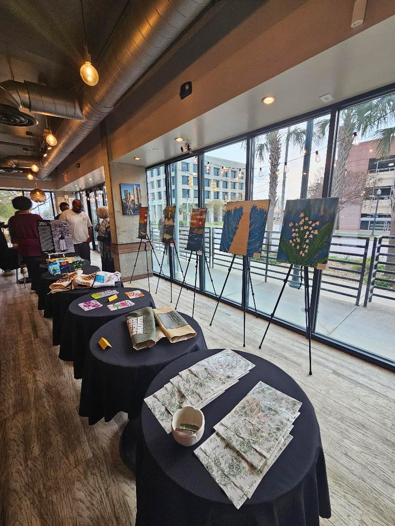 Gallery with paintings on easels and tables displaying art supplies inside a modern building with large windows.