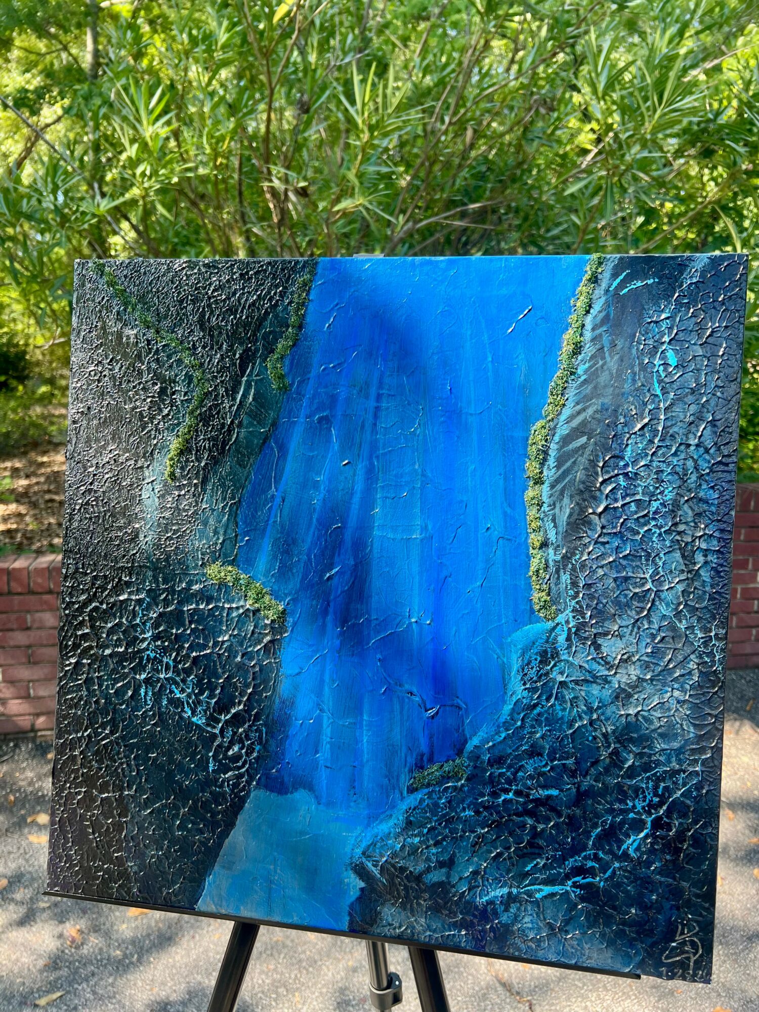 Abstract painting with blue, green, and black colors, depicting a waterfall or flowing water scene, on an easel outdoors.