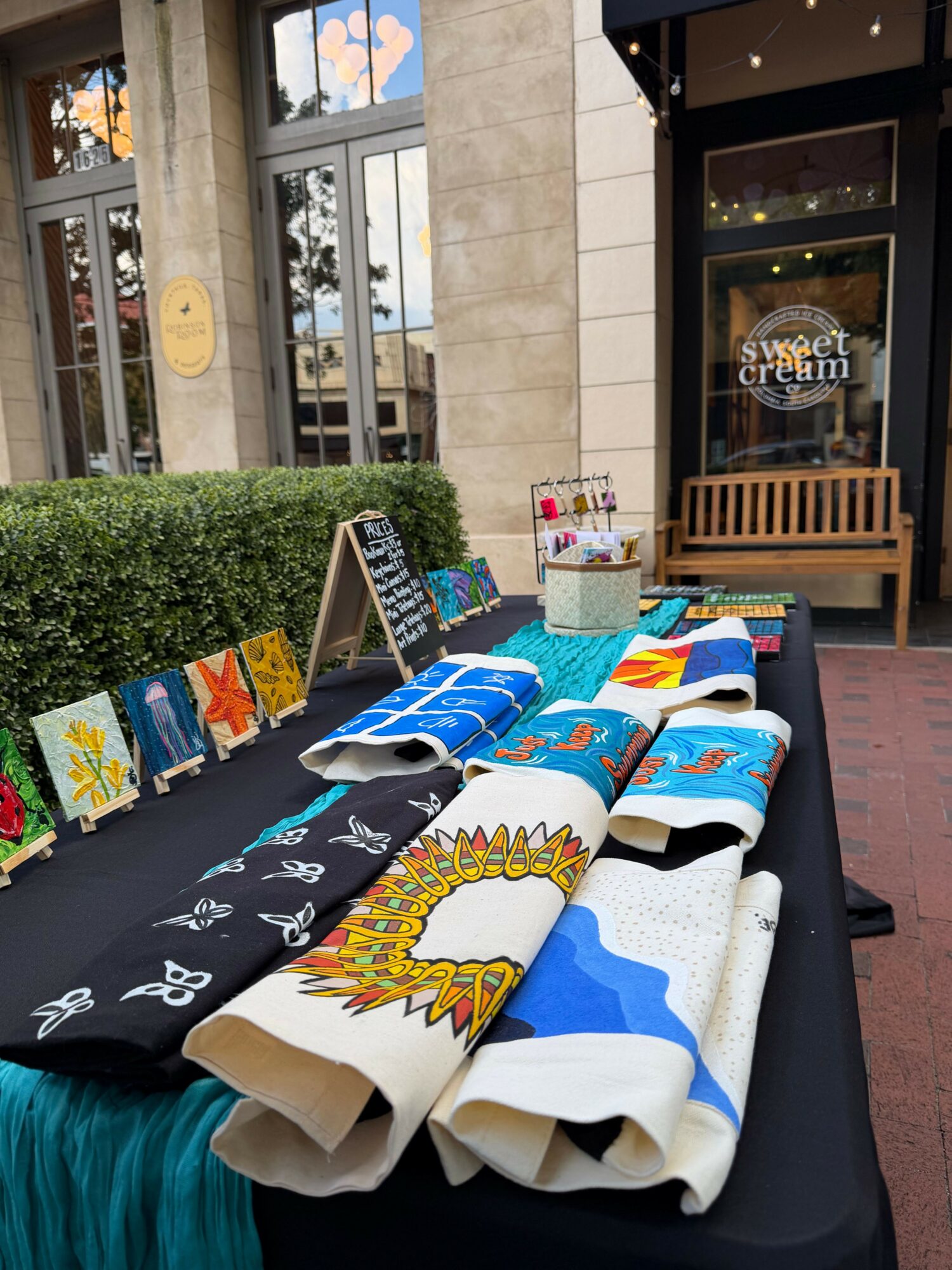 Table with colorful rolled fabrics and artwork displayed outside a building with glass doors and windows.