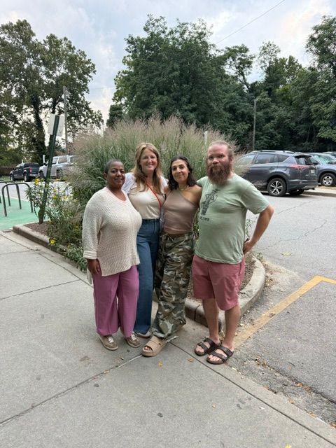 Four people standing on a sidewalk, with trees and parked cars in the background.