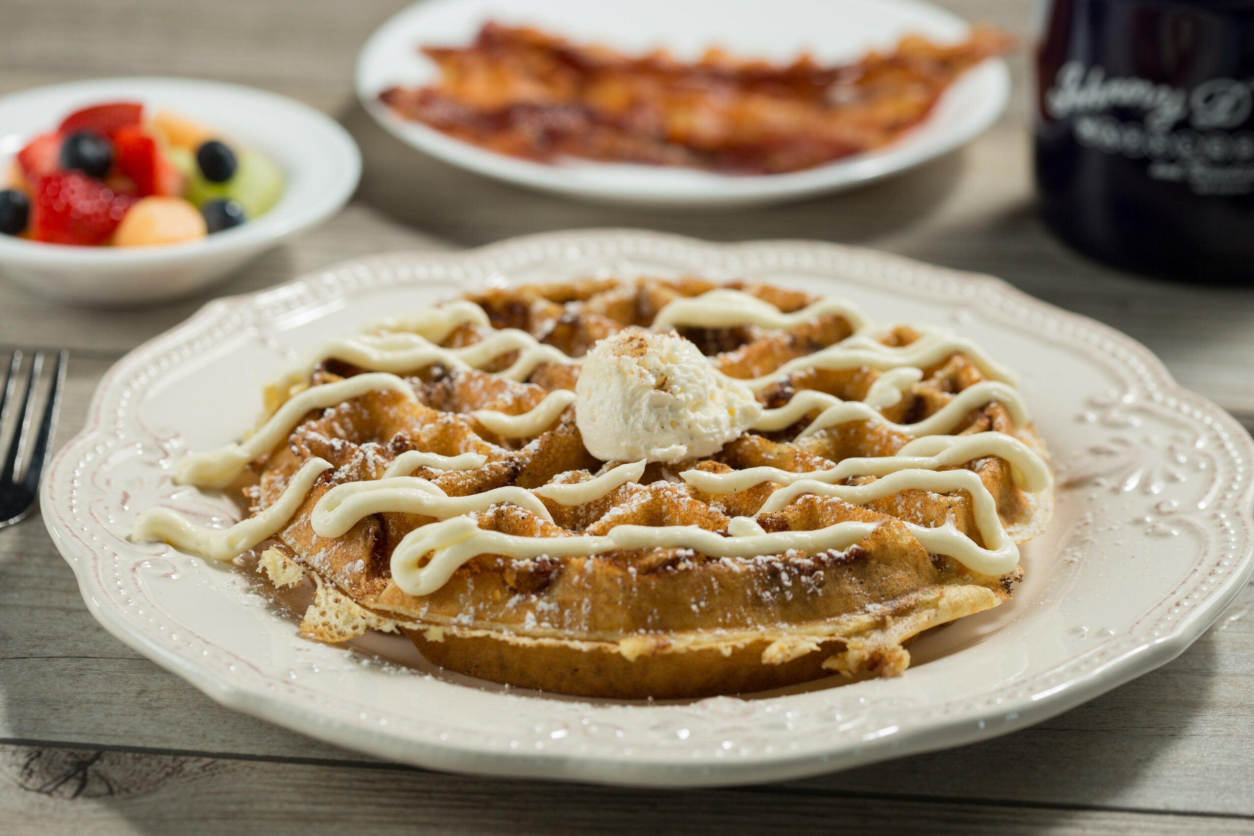 Waffle topped with whipped cream and drizzled with syrup on a decorative plate, with fruit, bacon, and a drink in the background.