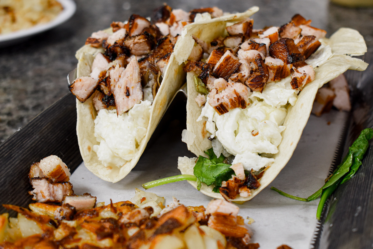 Two tacos filled with chopped meat and vegetables on a plate, garnished with cilantro, on a dark surface.
